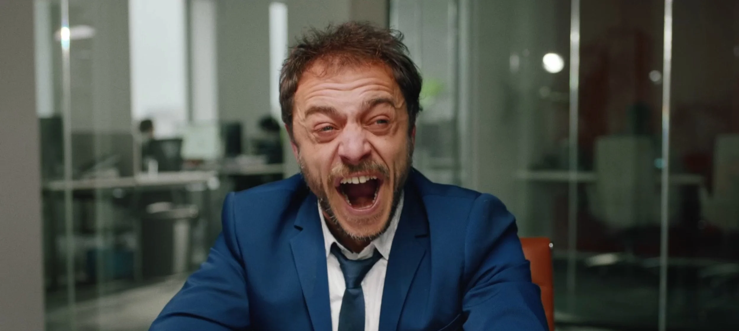 A man with disheveled hair and a beard, dressed in a blue suit and white shirt, is yelling with his mouth wide open and eyes squinted, sitting at a desk in an office setting.