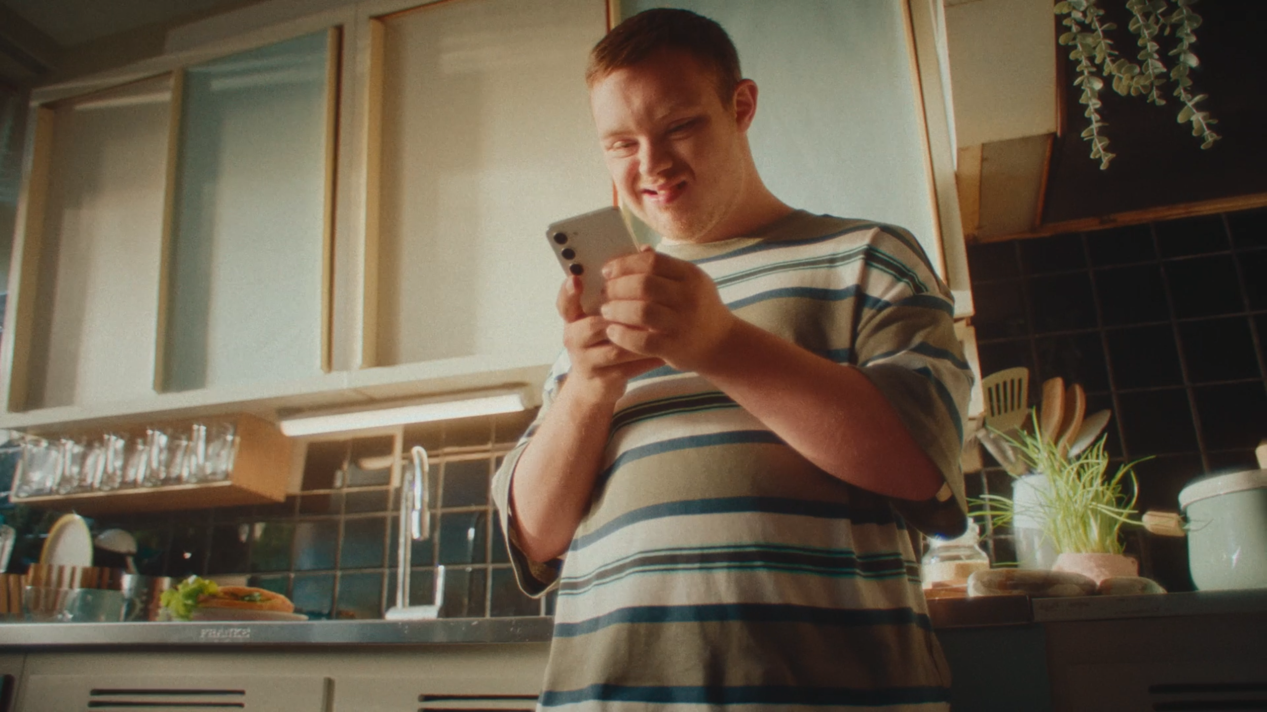 A young man with short red hair and a striped t-shirt is looking at his phone in a kitchen, smiling.