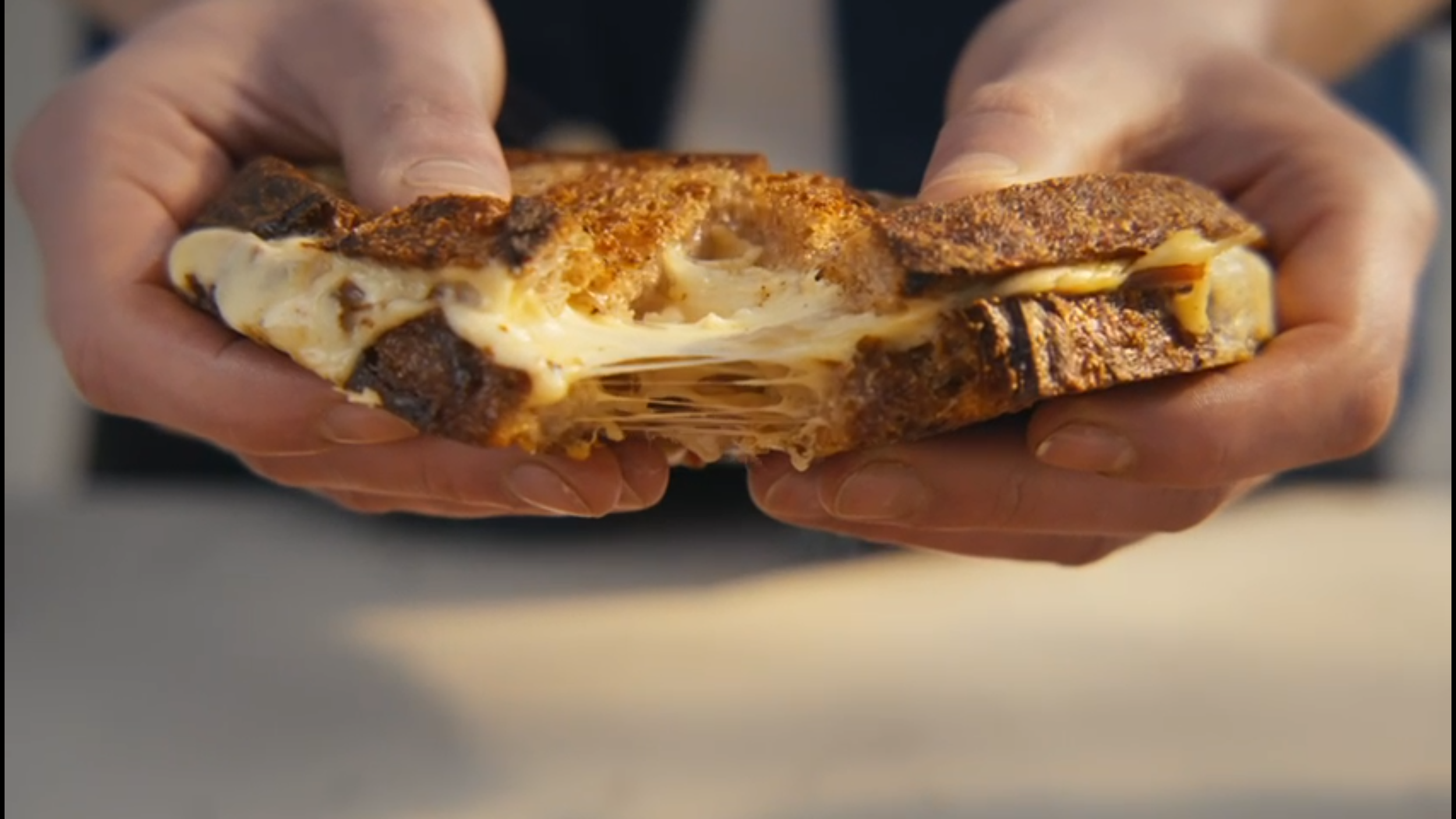 A person holding a large, toasted, and cheesy grilled cheese sandwich with melted cheese stringing from the middle.