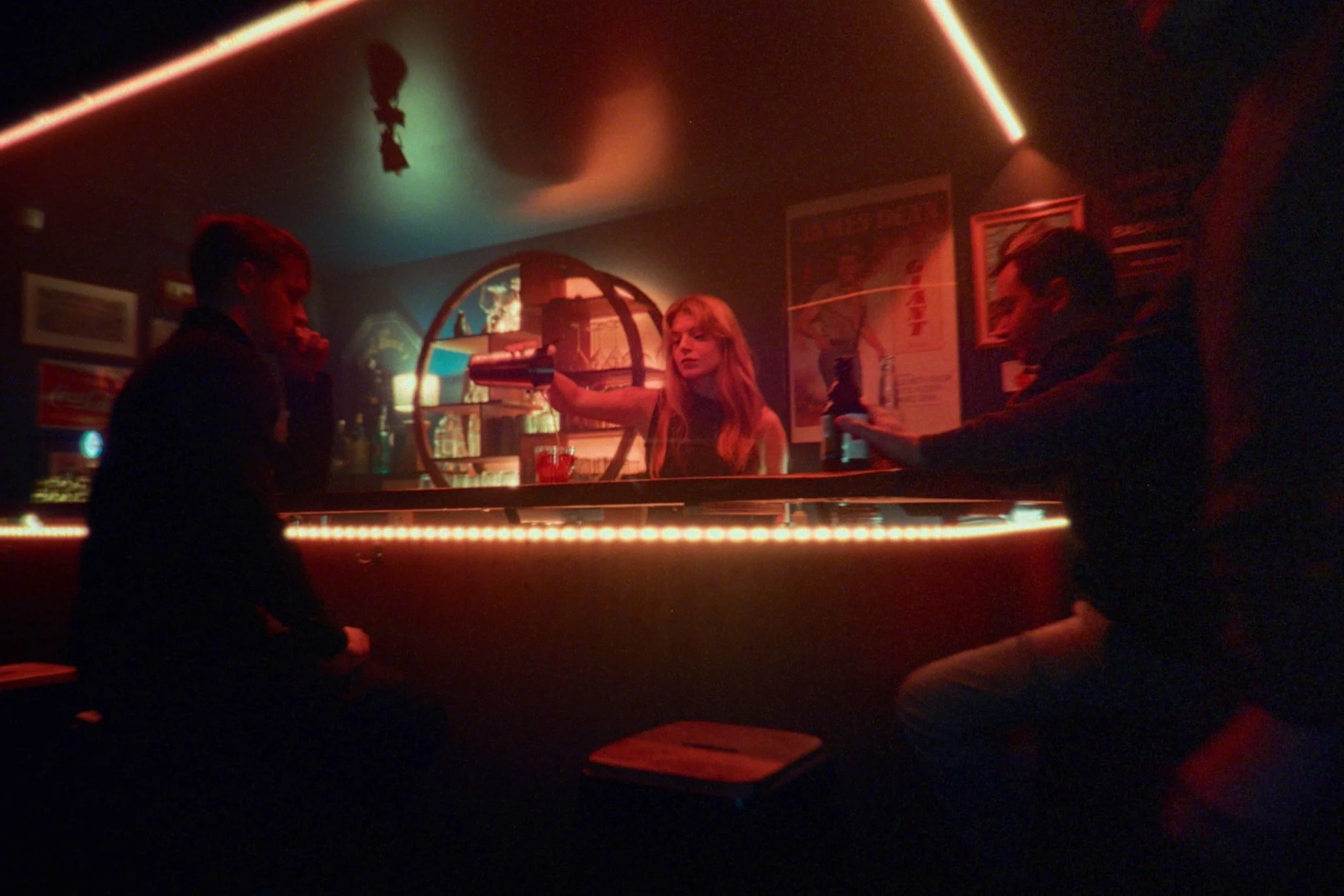 A dimly lit bar with a woman behind the counter serving drinks to two men, one on each side. The bar has a neon-lit counter and posters on the wall, creating a moody atmosphere.