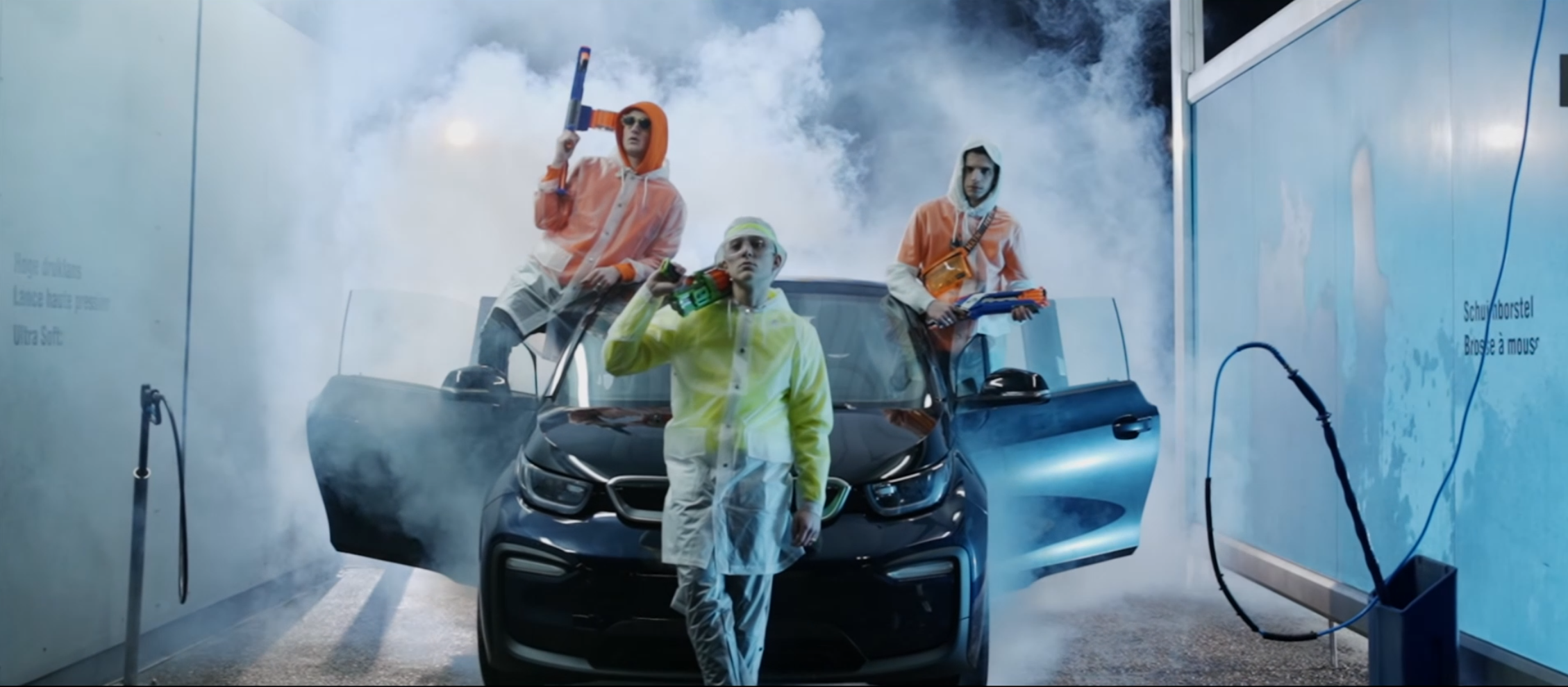 Three young men in colorful jackets and sunglasses washing a black car at a car wash station with steam and water spray around them.