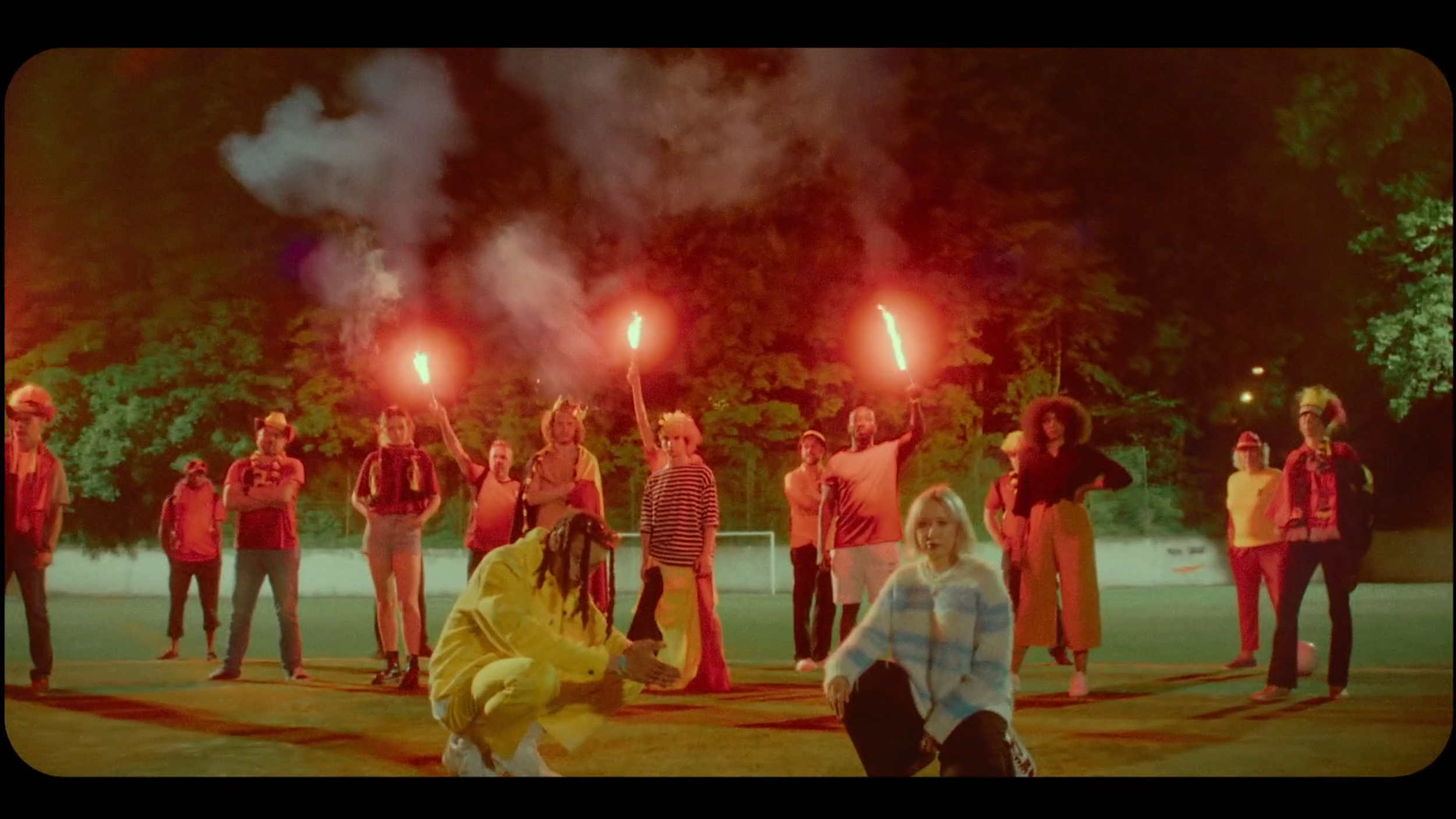 A group of diverse people in colorful attire, some holding flares with smoke and light, standing on a field at night with trees illuminated by green lighting in the background.