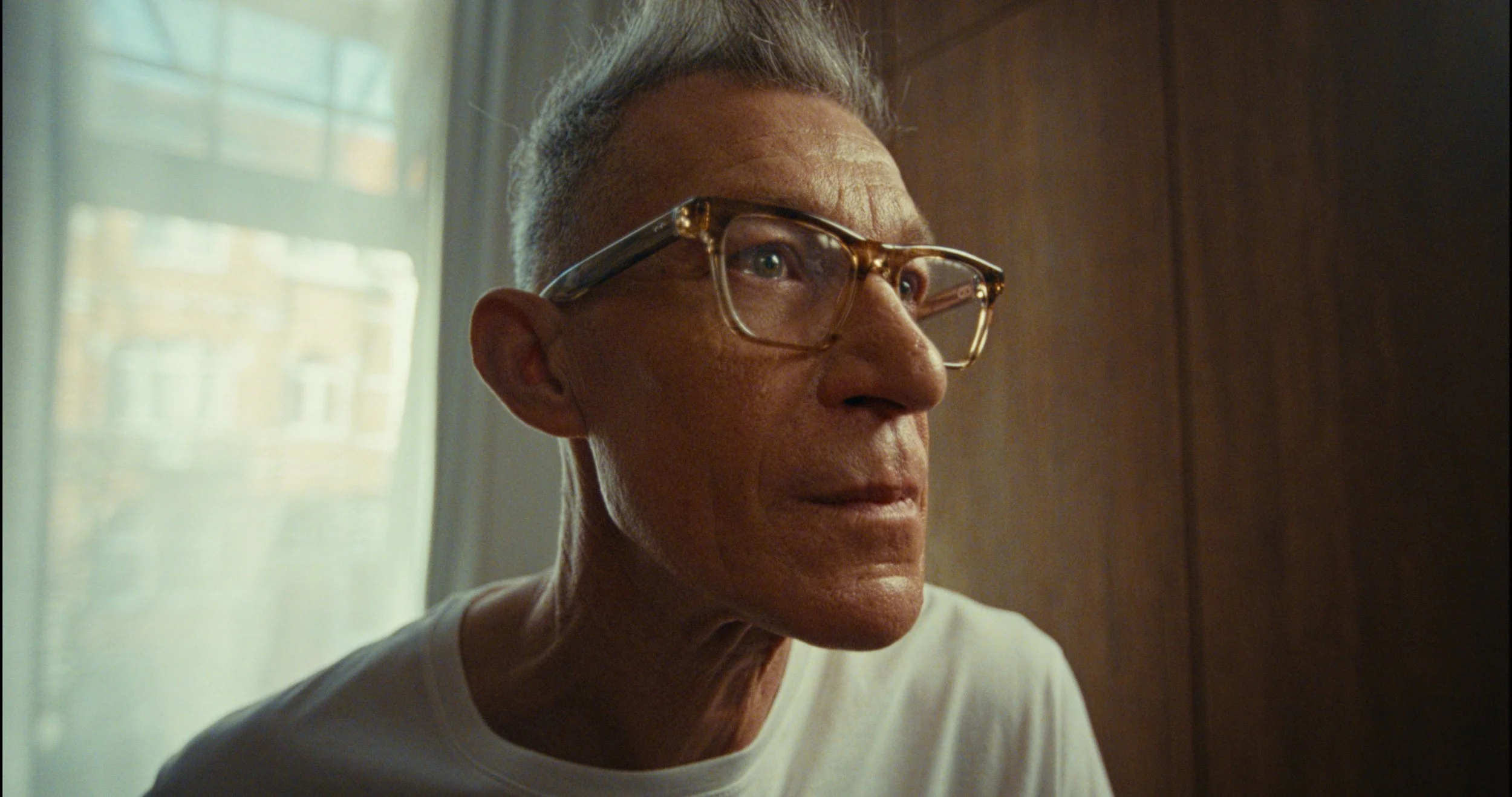 Close-up of an older man with glasses, looking thoughtfully to the side, indoors with natural light coming through a window.
