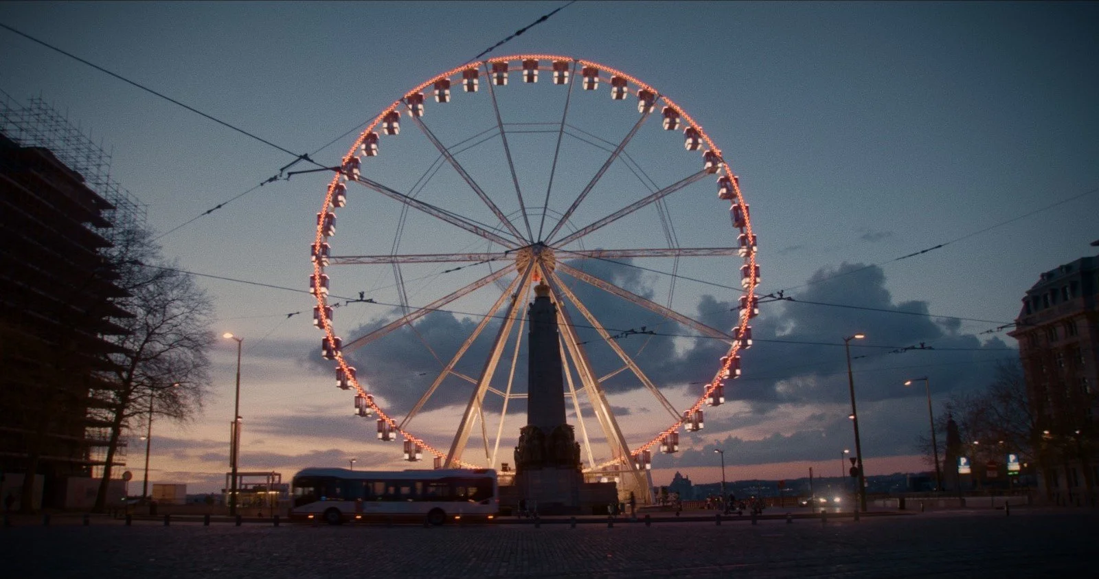 A large illuminated Ferris wheel at dusk with a bus passing by on the street below.