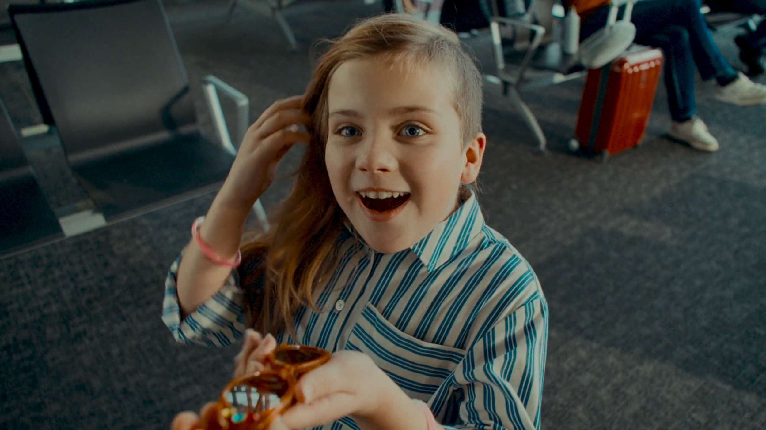 A young girl with long red hair, wearing a blue and white striped shirt, smiling excitedly as someone offers her a slice of pizza inside an airport terminal filled with chairs and travelers.