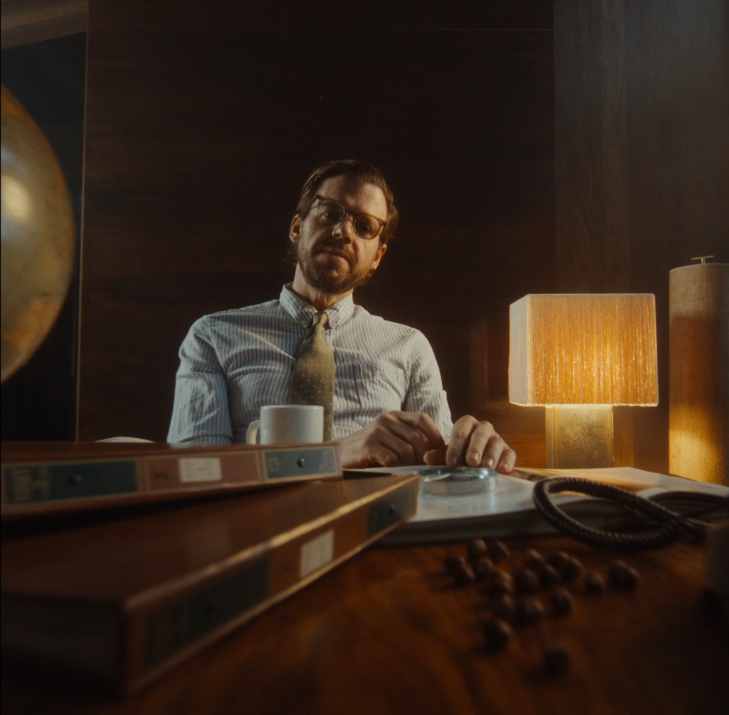Man with glasses sitting at a desk with a cup, a lamp, and a book, looking downward with a serious expression.