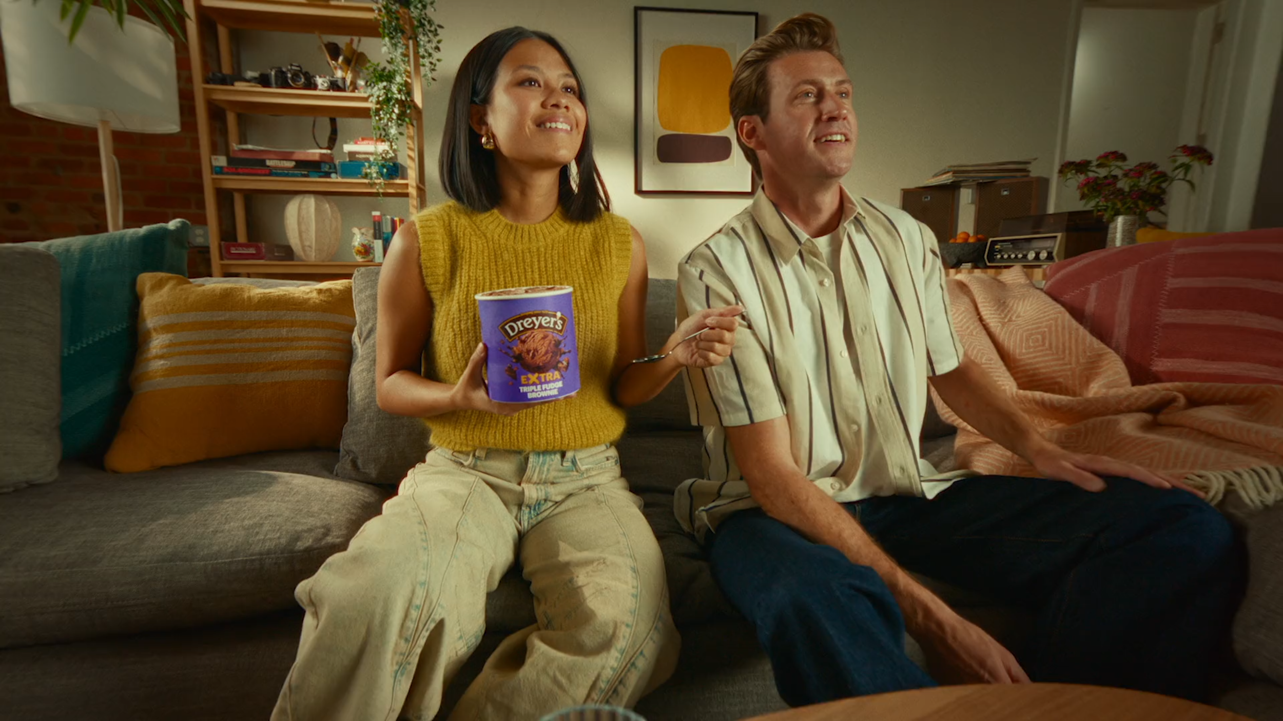 A woman with dark hair holding a tub of Dreyer's ice cream, sitting on a sofa next to a man in a striped shirt, both watching TV in a cozy living room.