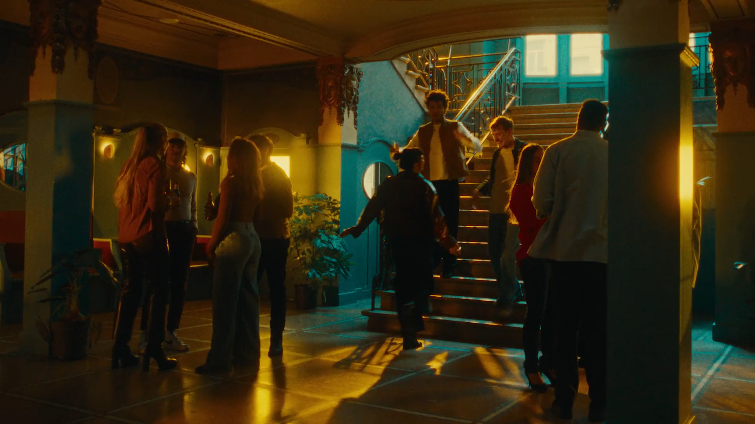 People dancing and socializing on a staircase and floor in a dimly lit venue, with warm yellow and teal lighting.