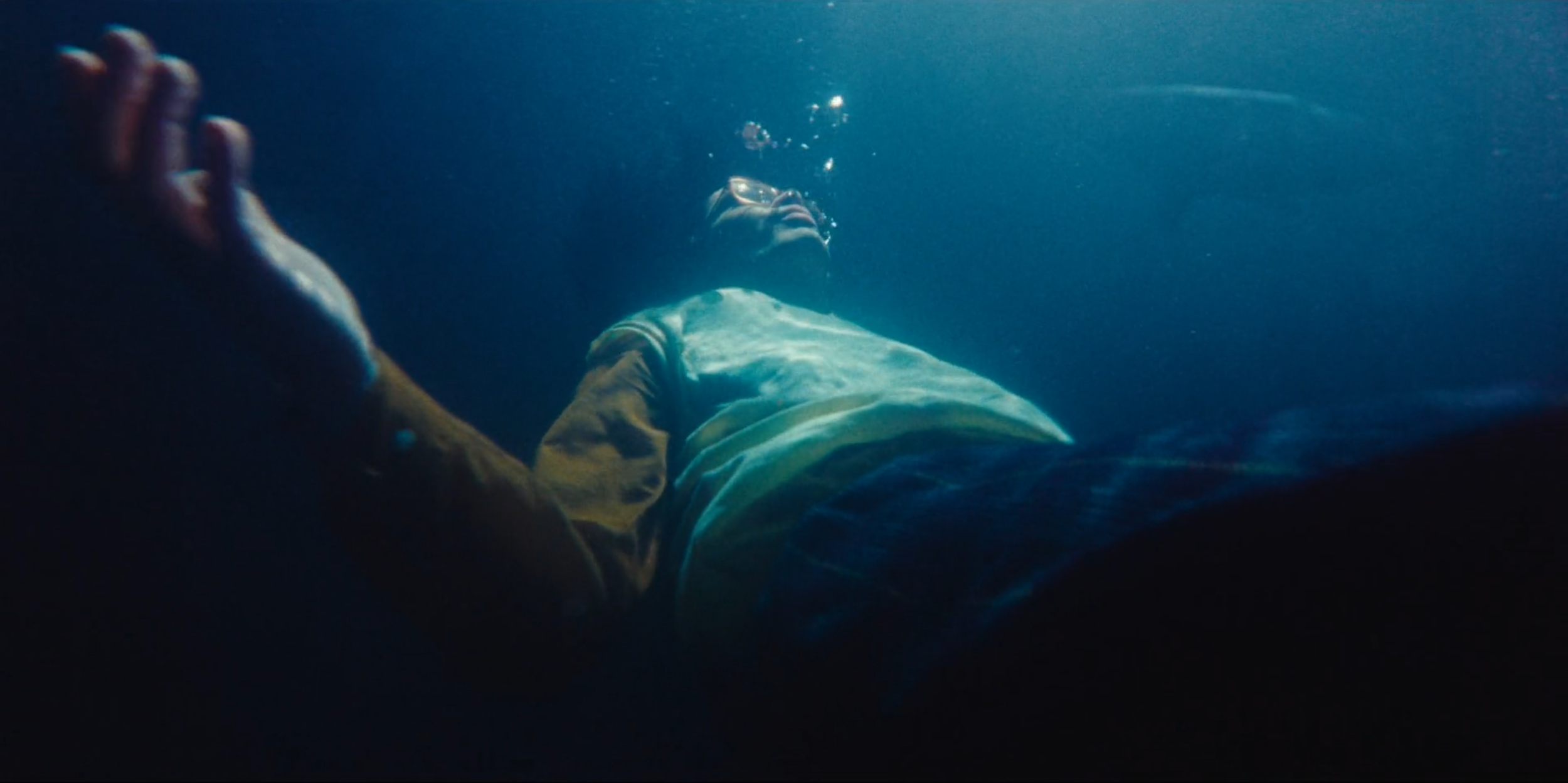 A person wearing glasses lying underwater with bubbles escaping from their face, their eyes closed and arms stretched out.