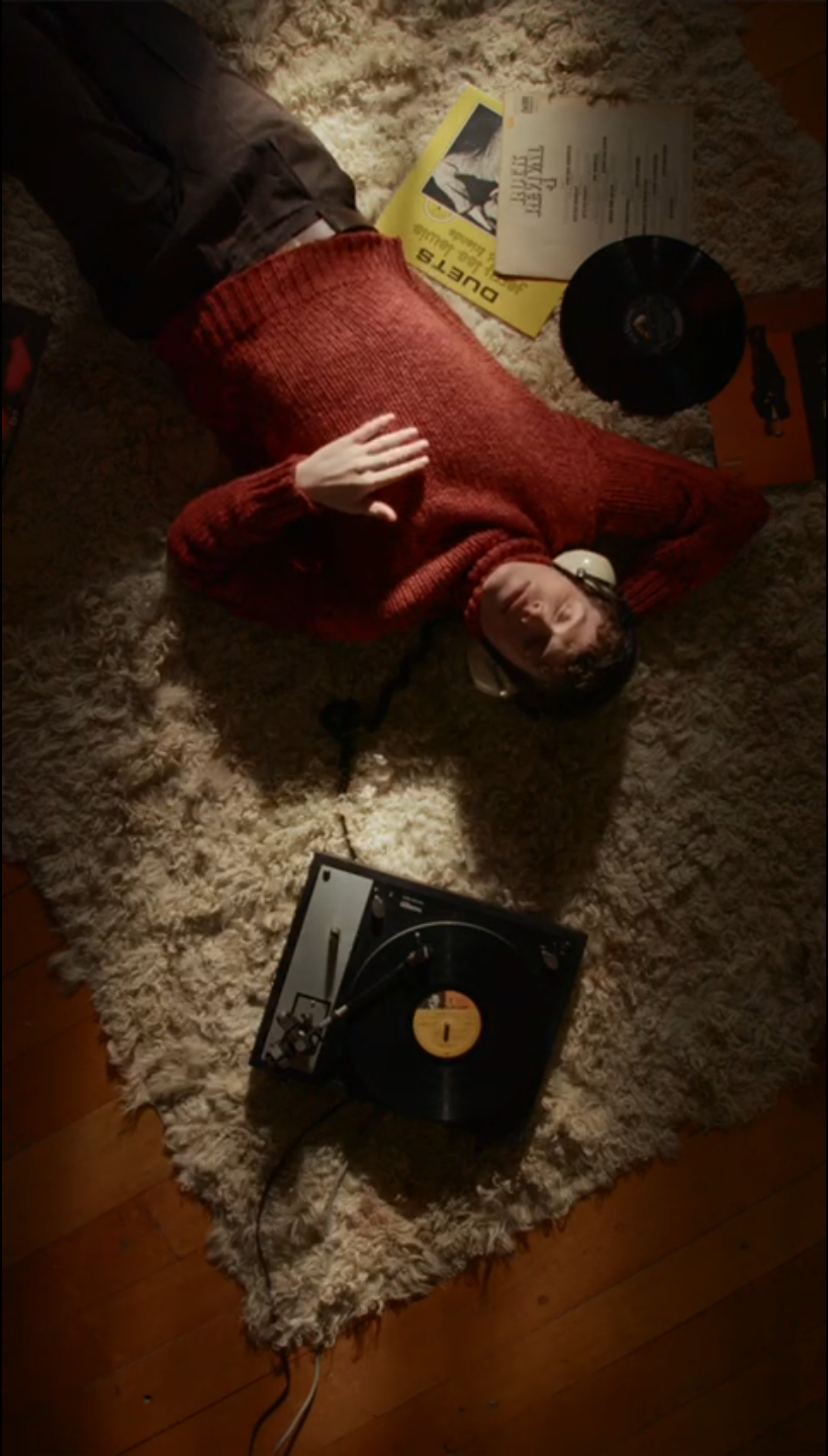 A person lying on a shaggy rug next to a turntable, with a yellow music album, a vinyl record, and some papers nearby. The person is wearing a red sweater and headphones.