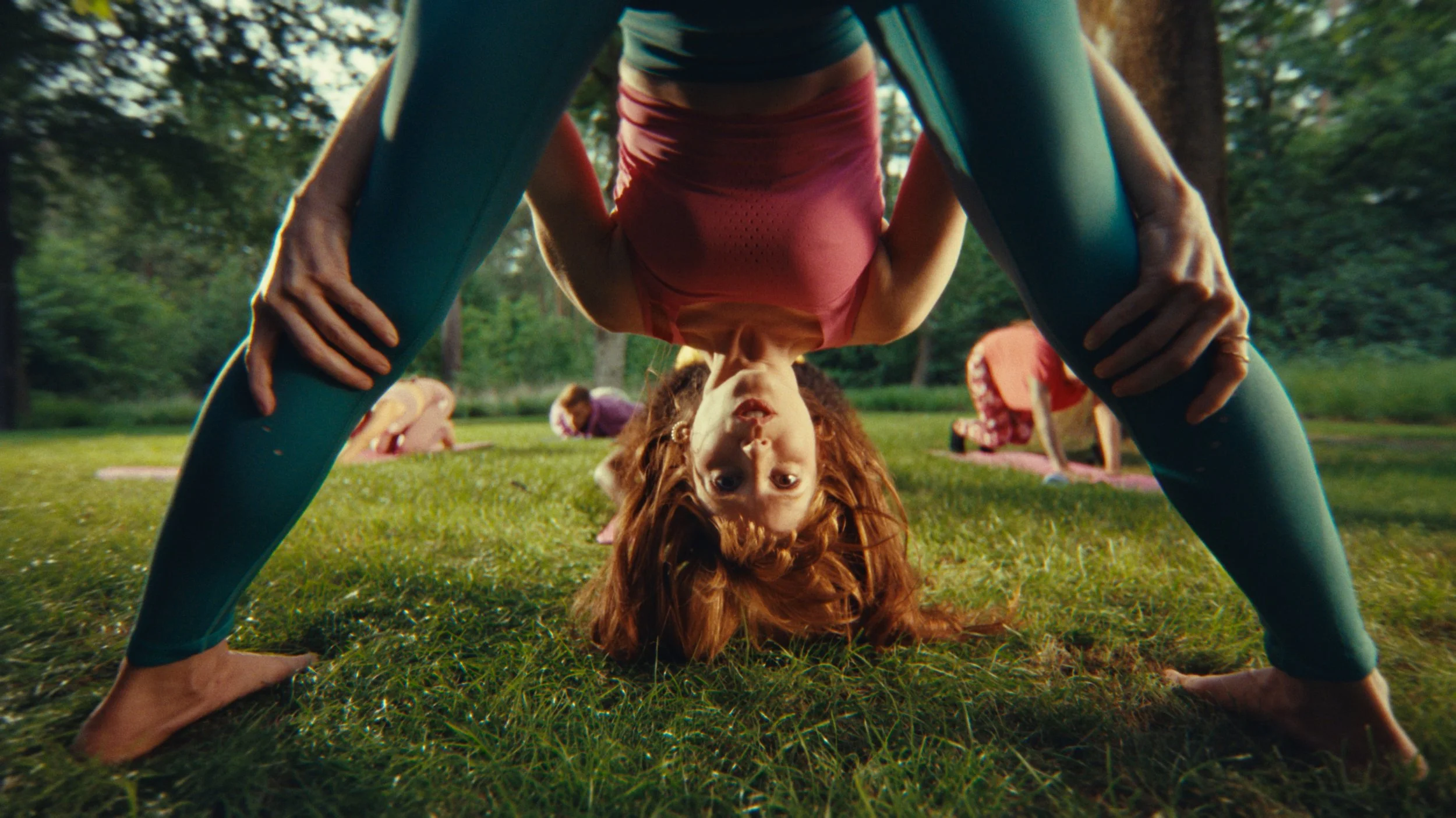 A woman in a yoga pose outdoors in a park during sunset, with other women practicing yoga in the background.