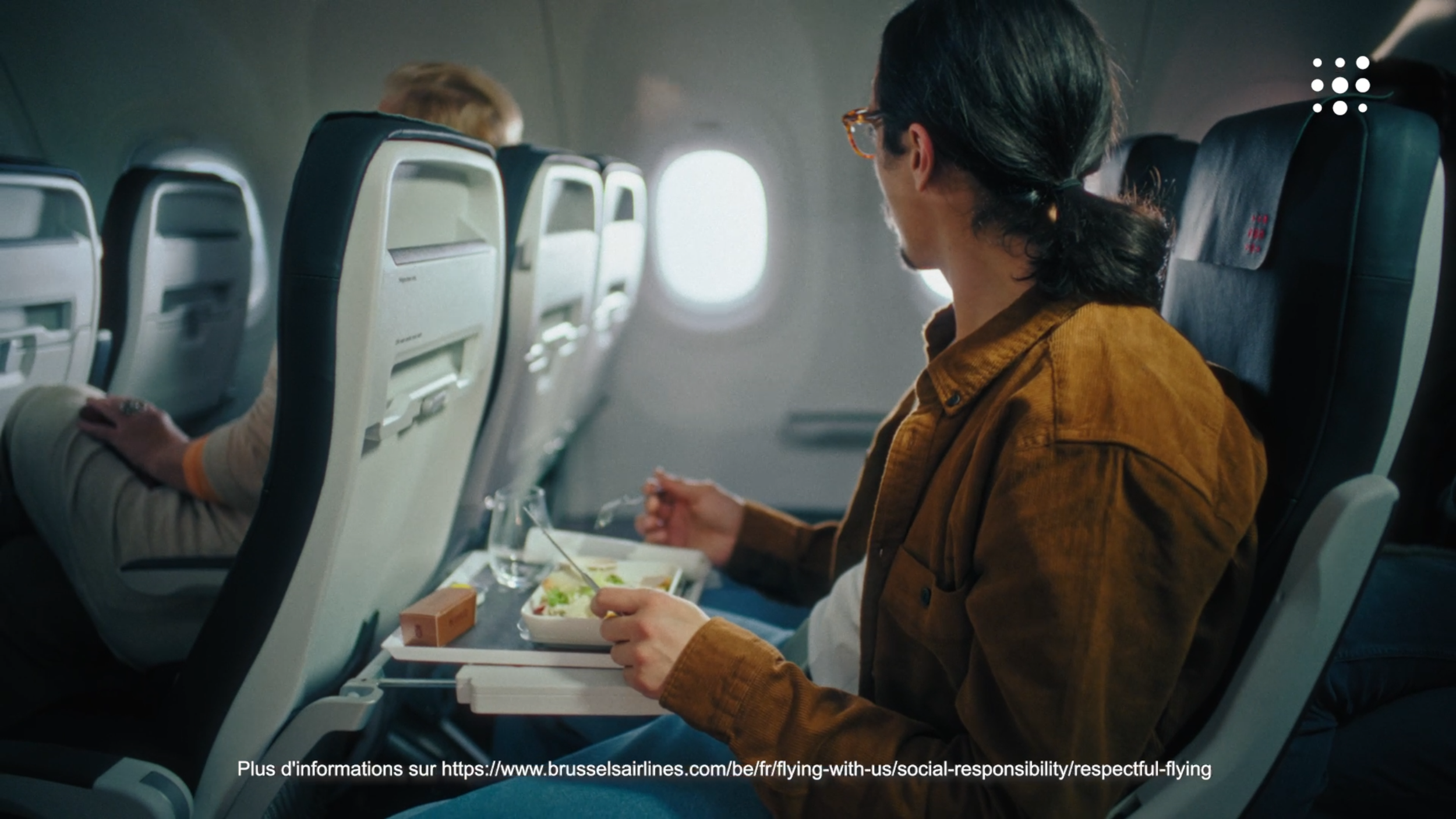 Passenger on an airplane eating a meal from a tray table, with other airplane seats and windows visible in the background.
