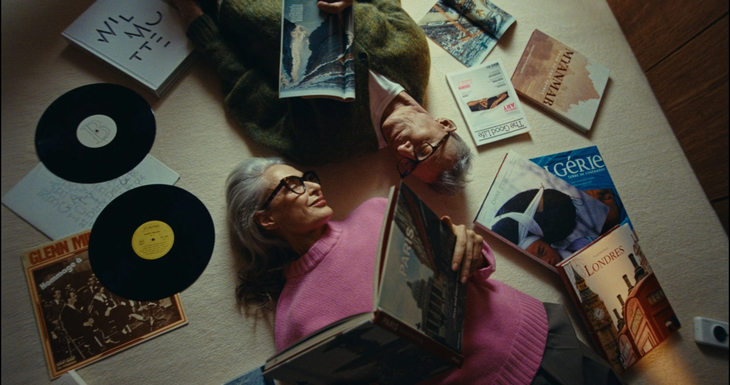 Two women lying on the floor surrounded by books, magazines, vinyl records, and a camera, engaging in reading and relaxing.