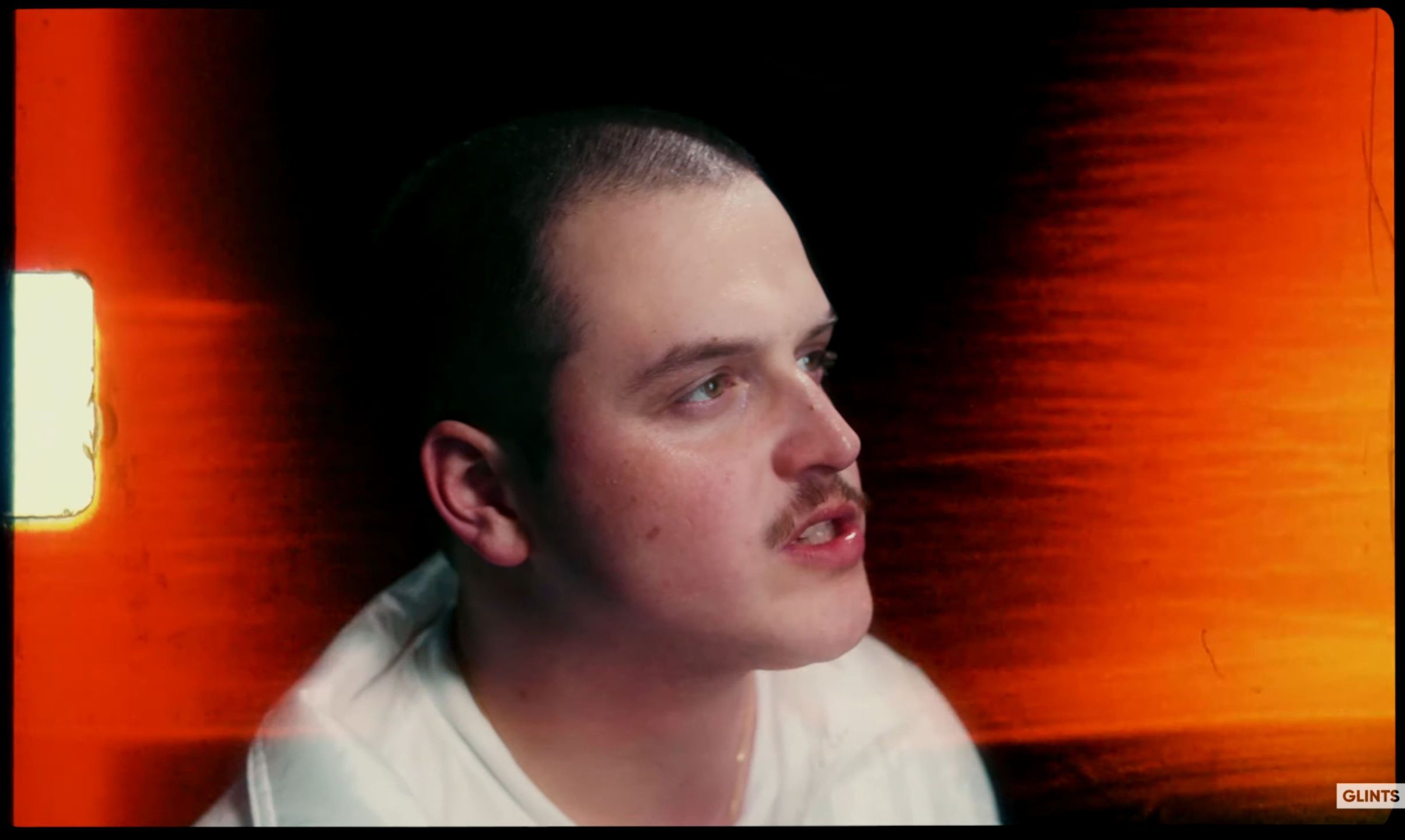 A young man with short dark hair and light skin, wearing a white shirt, sitting against a dark and orange background, looking to the right.