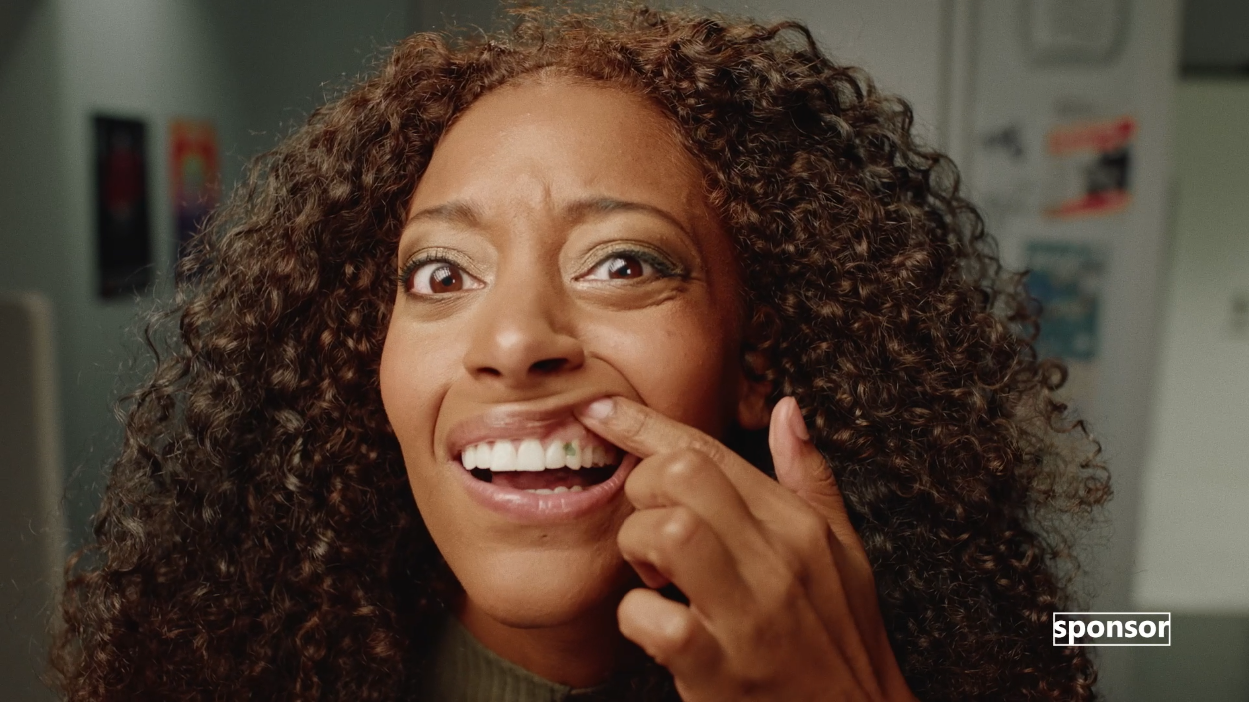 A woman with curly hair pulling a green piece of food from her mouth and smiling.