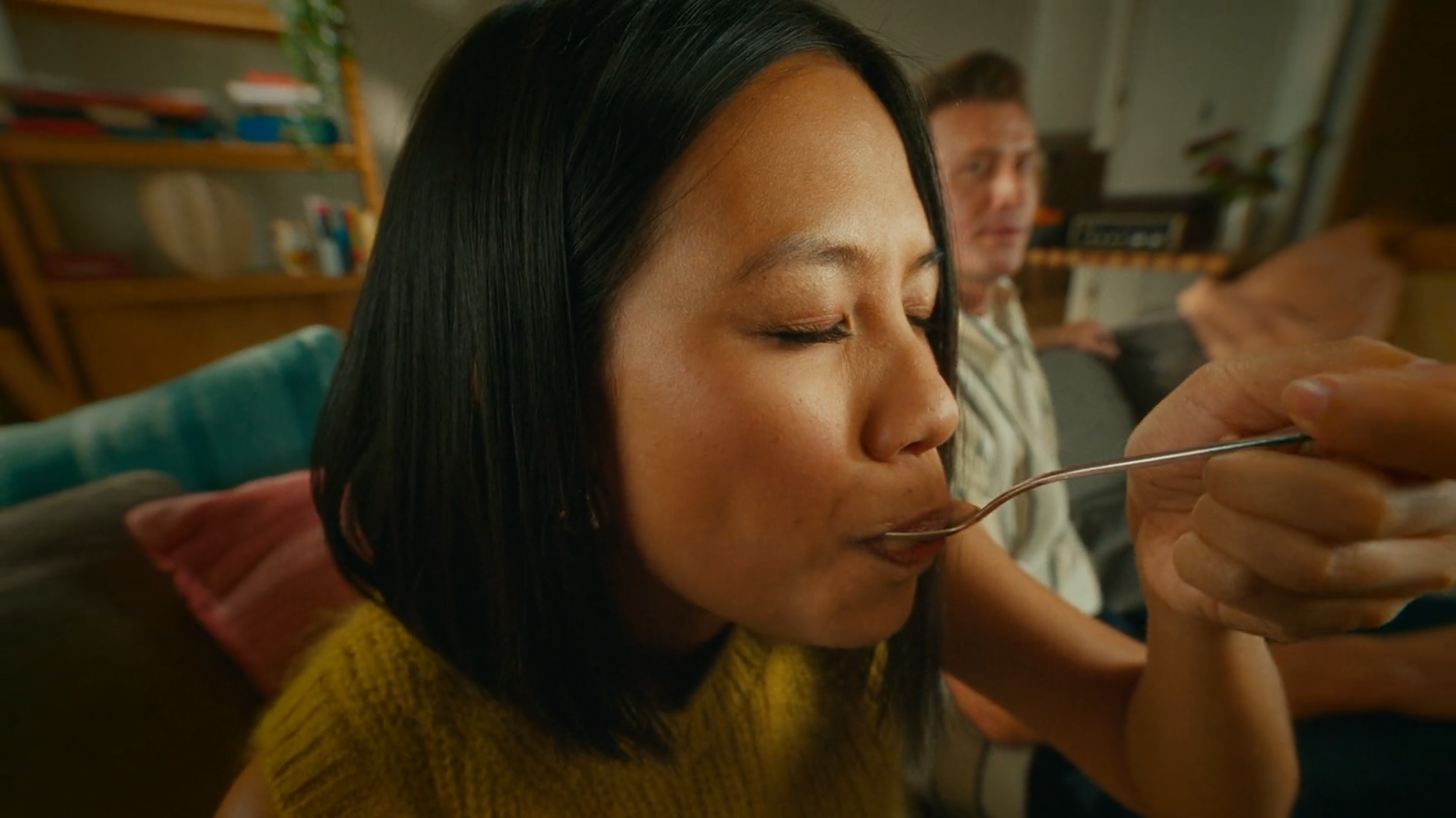 A woman with black hair is about to eat food with a spoon, with a man sitting in the background on a sofa in a cozy room.