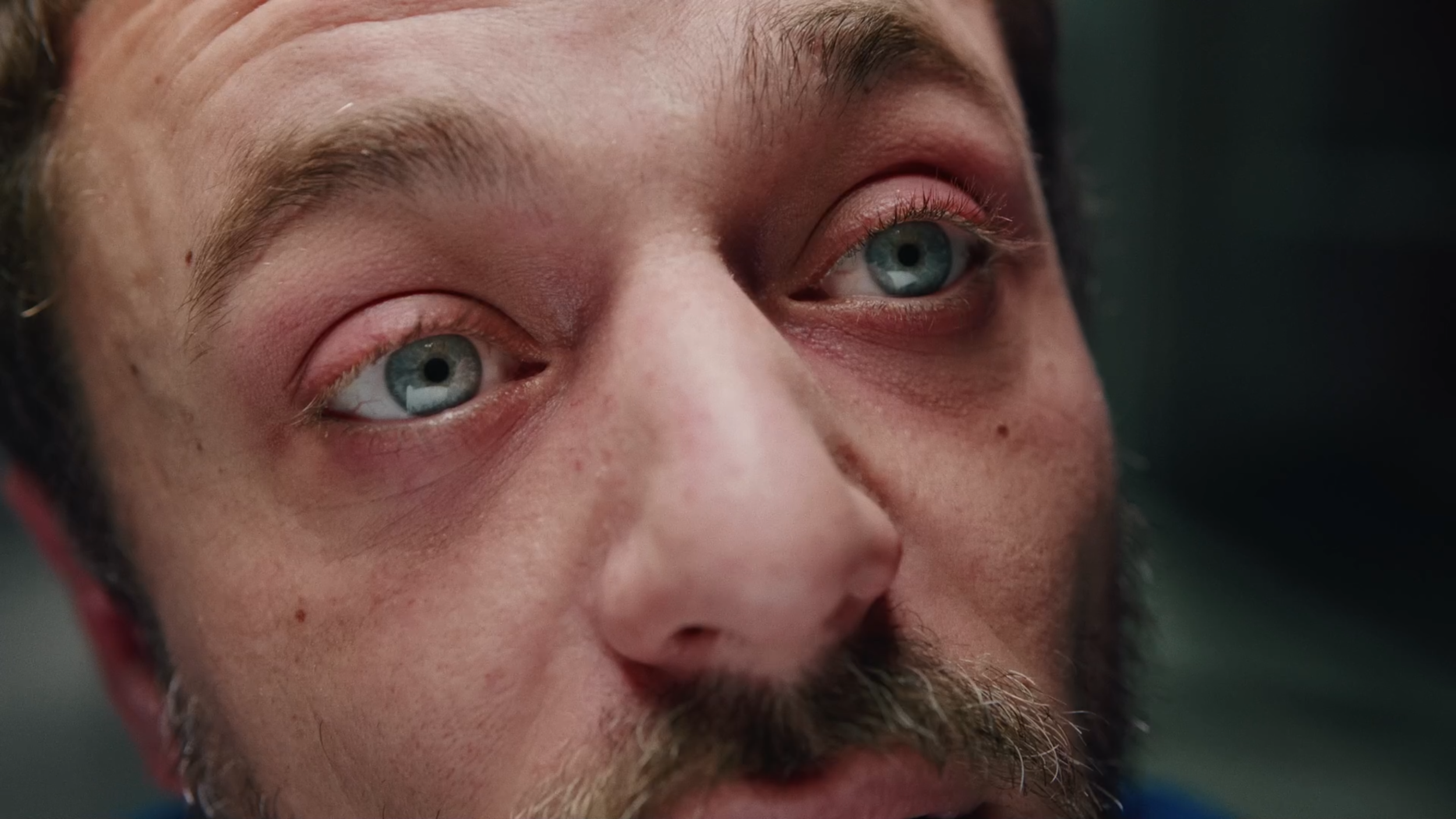 Close-up of a man's face, focusing on his blue eyes, with visible skin texture, facial hair, and dark hair.