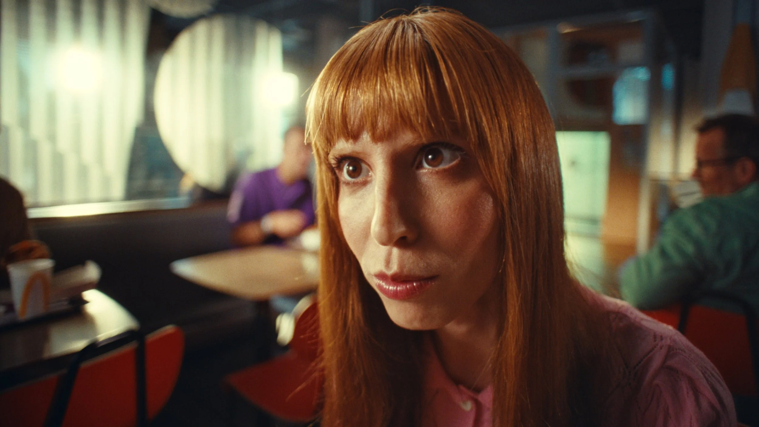 Close-up of a woman with long red hair and light makeup in a colorful cafe or diner setting, with blurred background of people and decor.