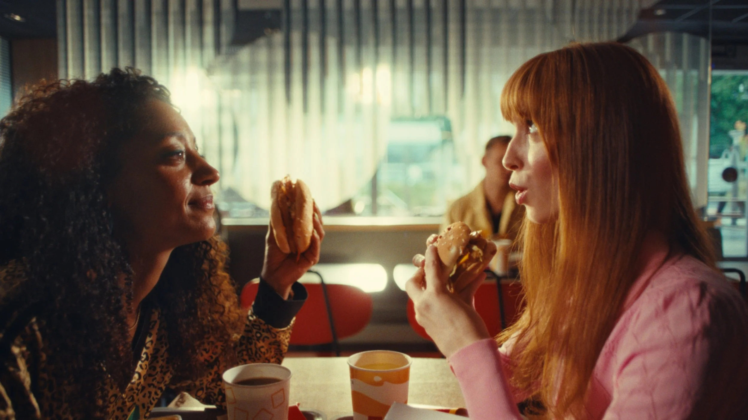 Two women having a conversation while eating fast food in a restaurant, showing close-up of their faces and burgers, with a window in the background.