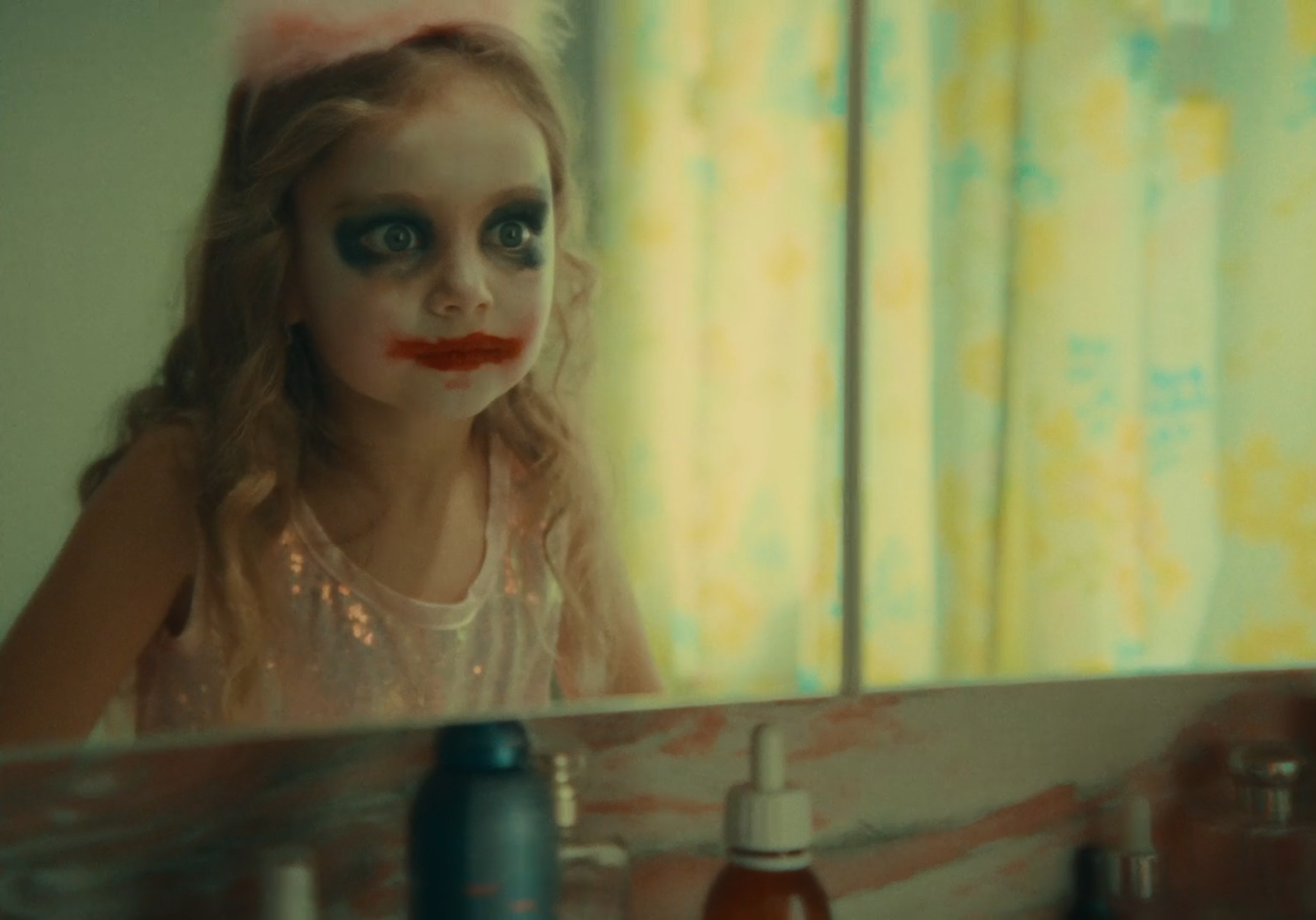 A young girl with makeup resembling a clown or Joker, with exaggerated red lips and dark eye makeup, looking into a mirror in a dimly lit room with patterned curtains in the background.