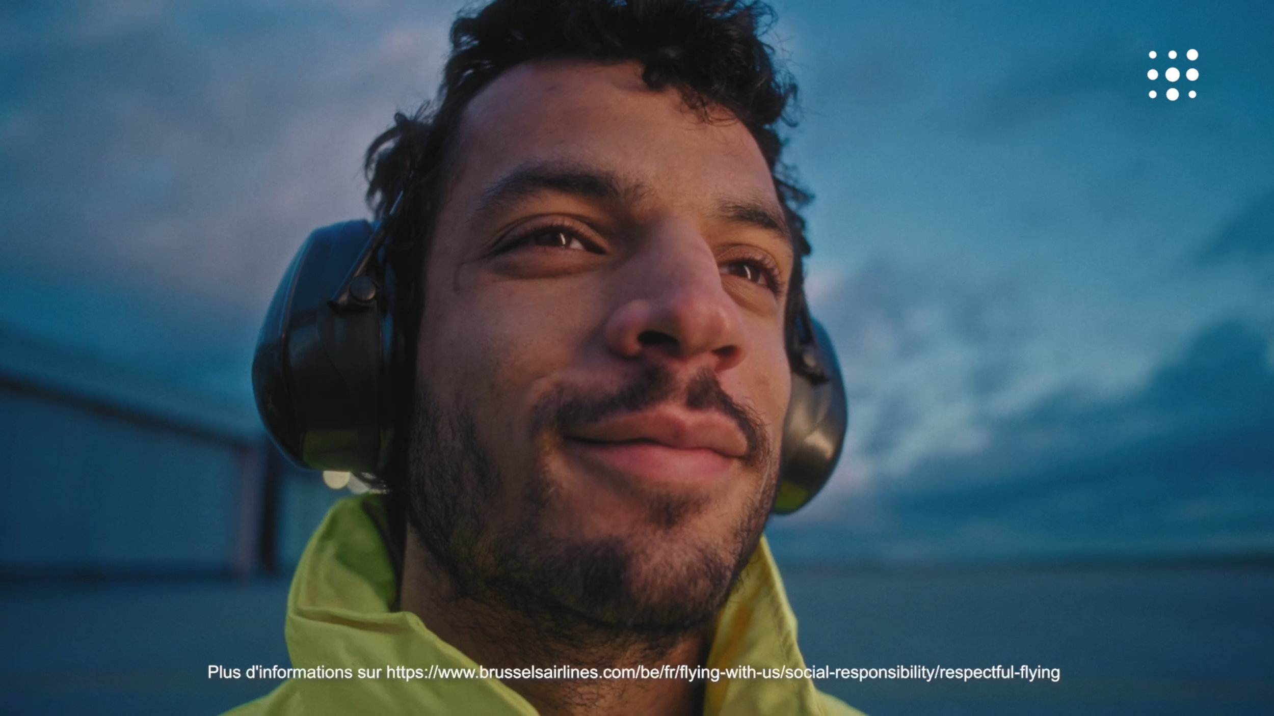 Close-up of a smiling man wearing headphones and a yellow jacket, with a cloudy sky background.