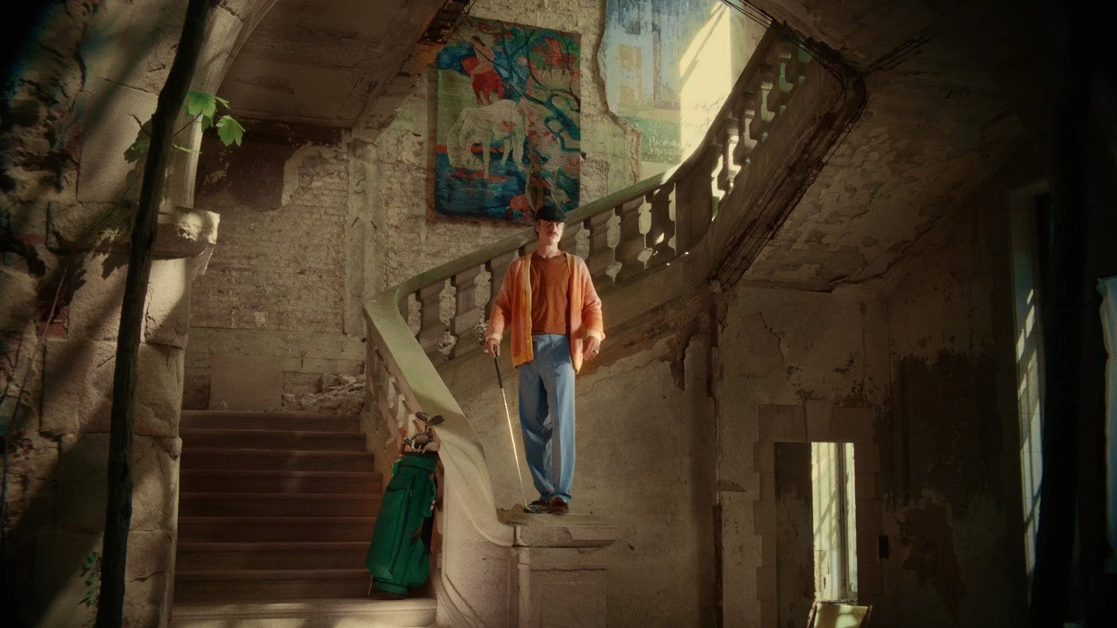 A young man in vintage golf attire standing on a worn staircase inside an old, dilapidated building with peeling walls, holding a golf club, with a golf bag beside him.