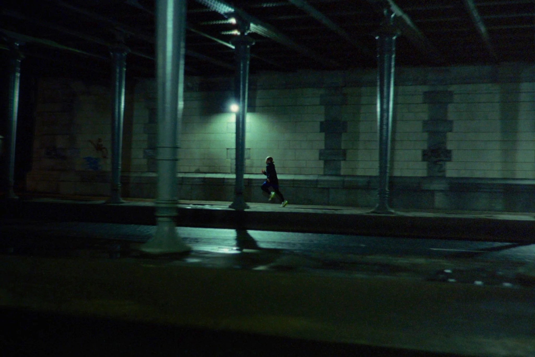 A person running on a sidewalk beneath a bridge at night, illuminated by a streetlight.