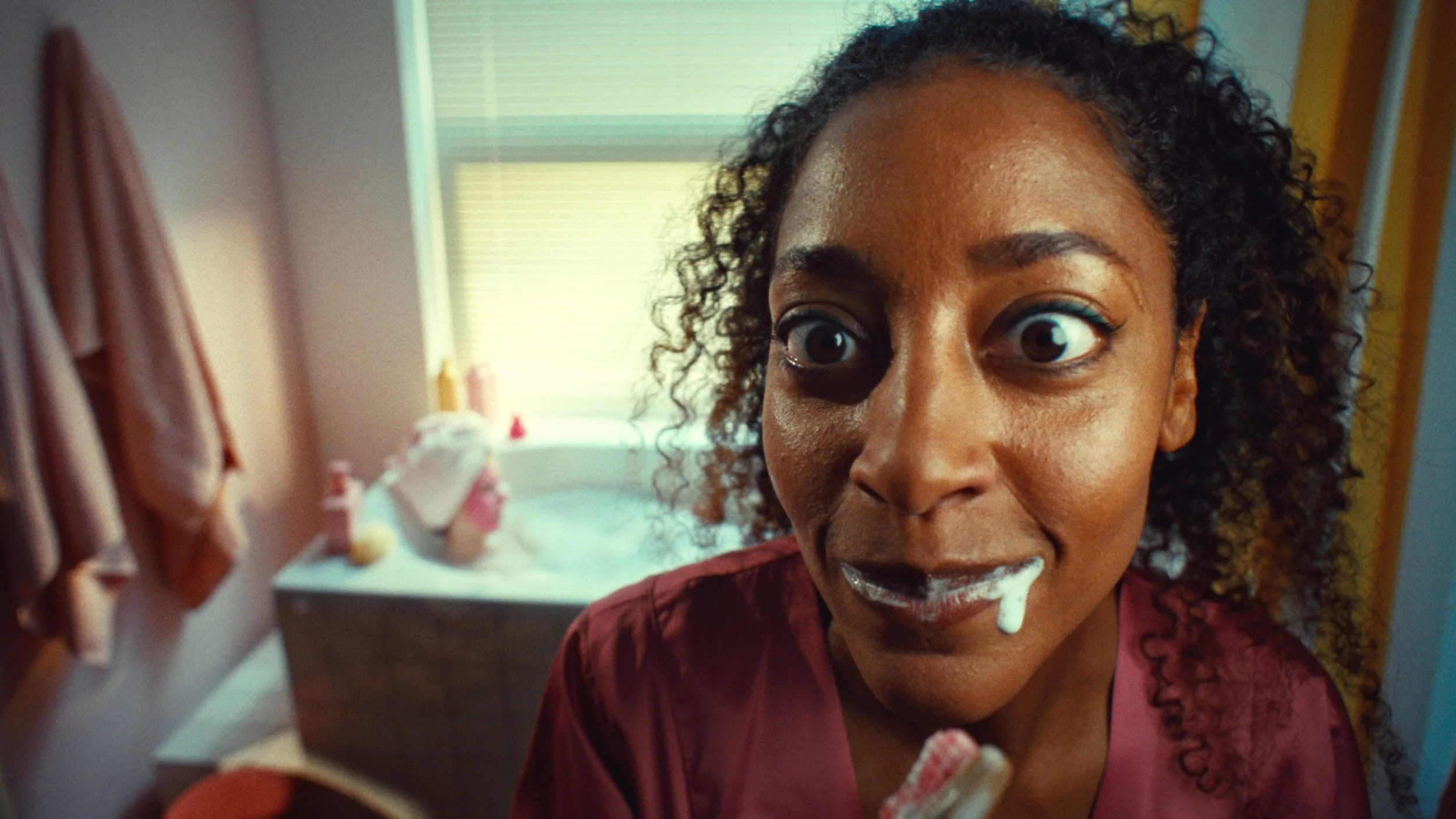 A woman with curly hair has foam on her lips from lathering soap, and she is looking excitedly at the camera while getting ready in a bathroom with a bathtub, towels on hooks, and a window with blinds in the background.