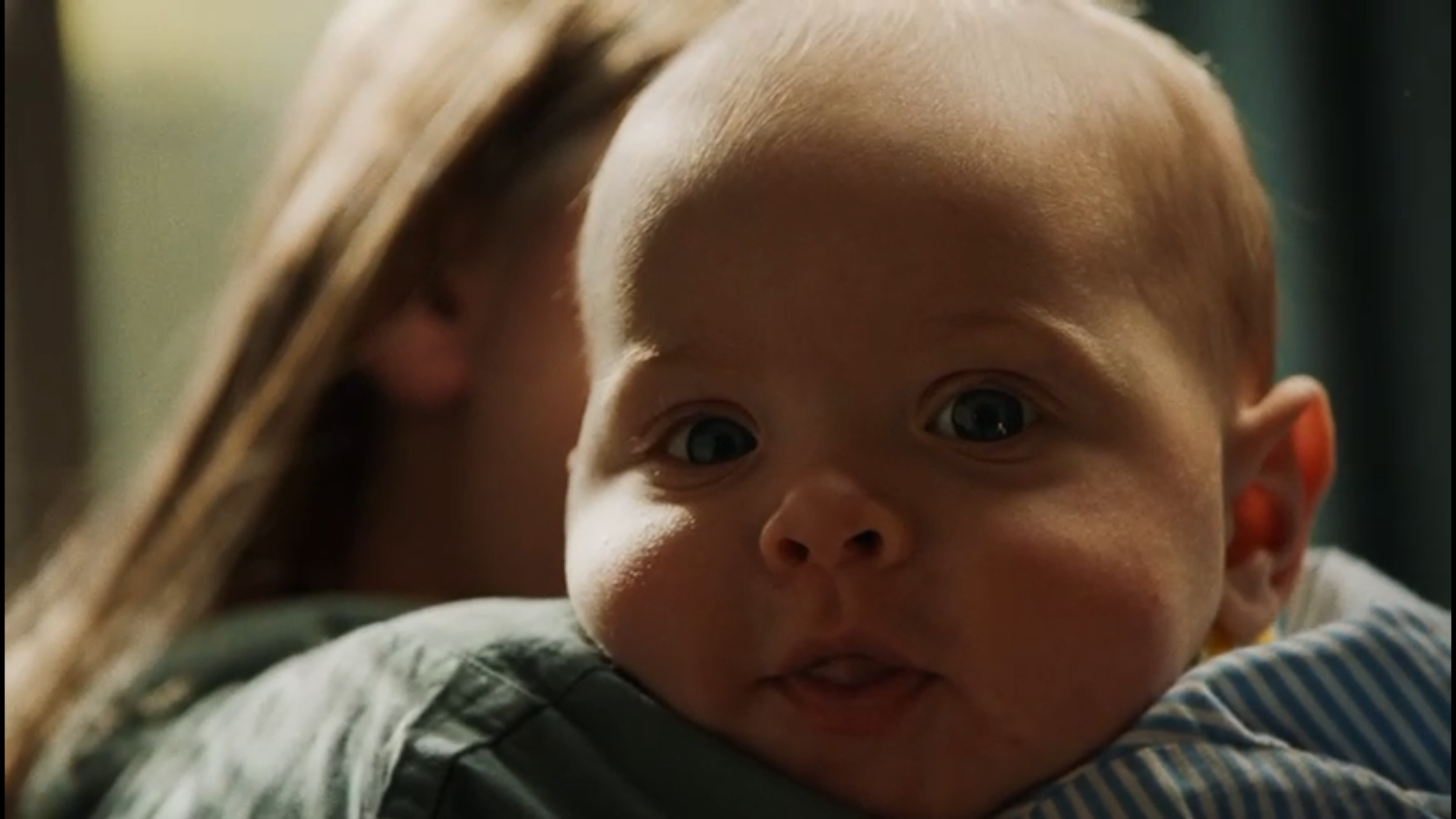 Close-up of a baby with light brown hair and big blue eyes, looking directly at the camera with a curious expression.