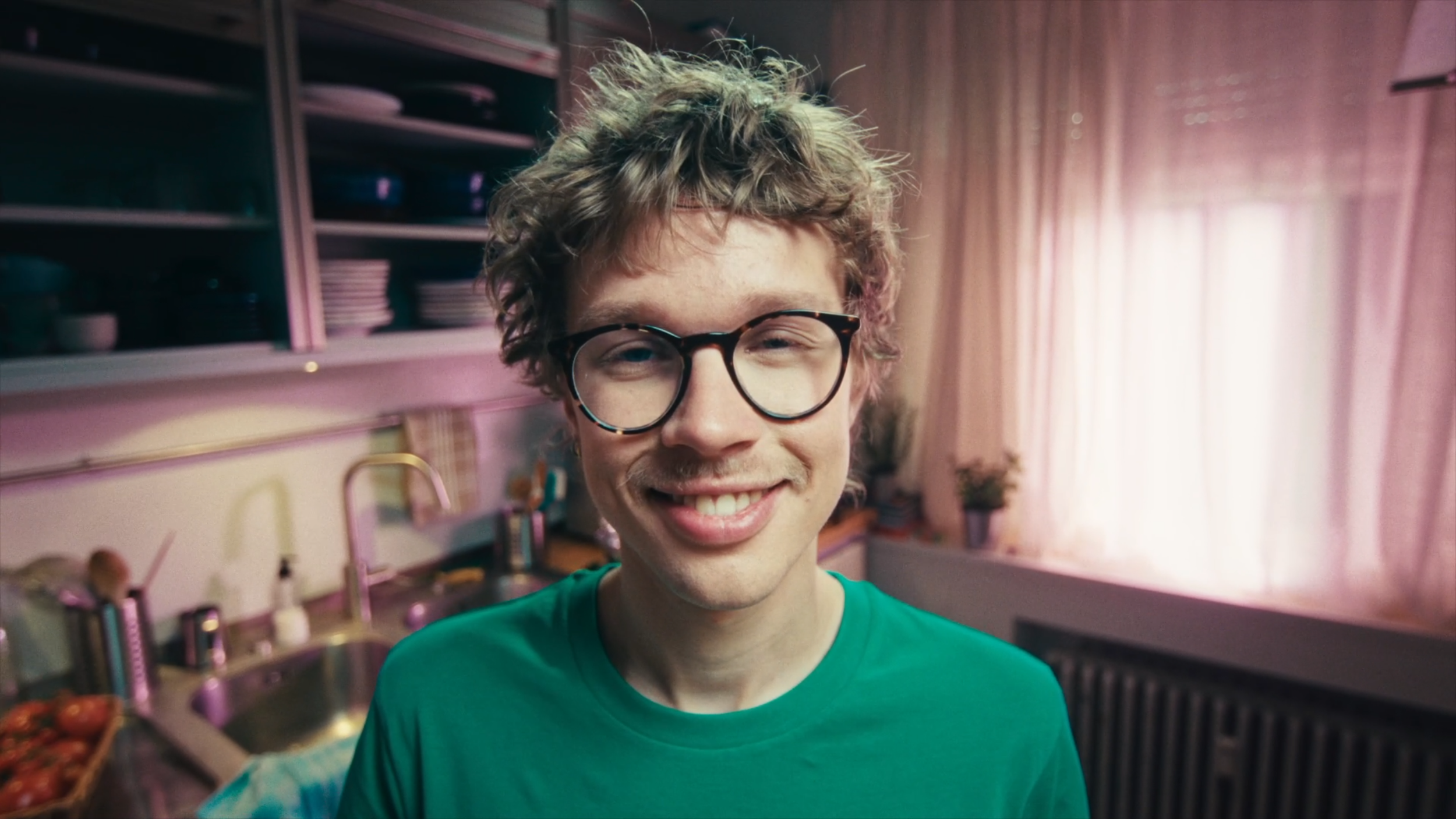 A smiling young man with curly hair and glasses in a pastel pink kitchen with open shelves and a window with pink curtains