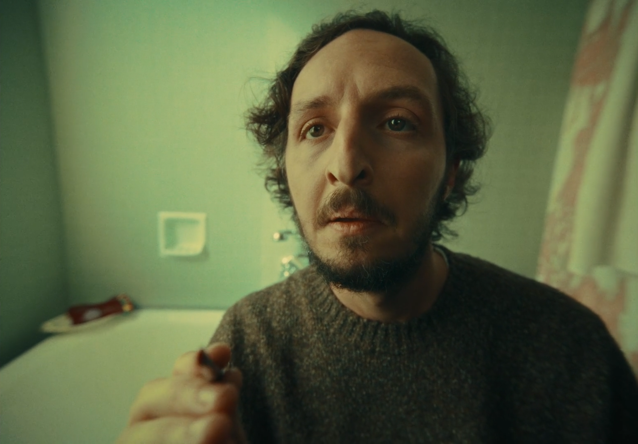 Close-up of a man with curly hair and beard, wearing a brown sweater, looking at the camera in a room with green walls and a bed in the background.