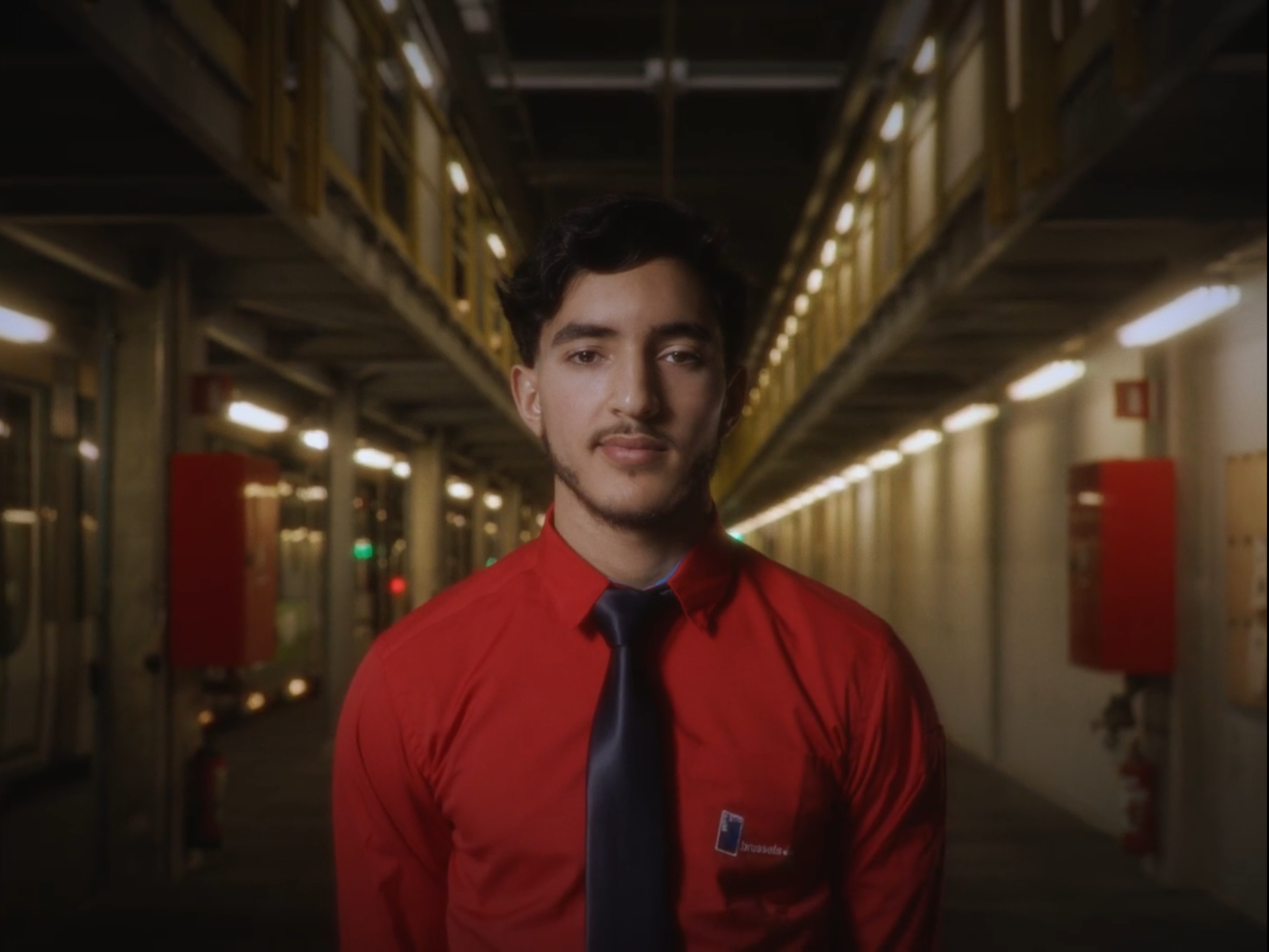 A young man wearing a red shirt and black tie standing in an industrial or underground setting with yellow railings and bright lights.