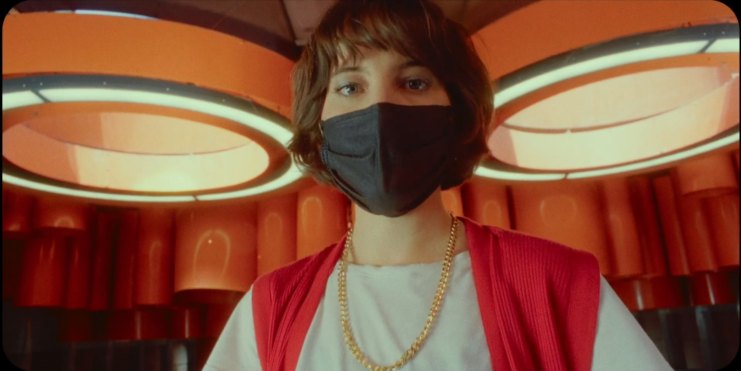 A person with shoulder-length brown hair wearing a black face mask, white T-shirt, red vest, and gold chain necklace, standing inside a modern building with orange circular lighting fixtures on the ceiling.