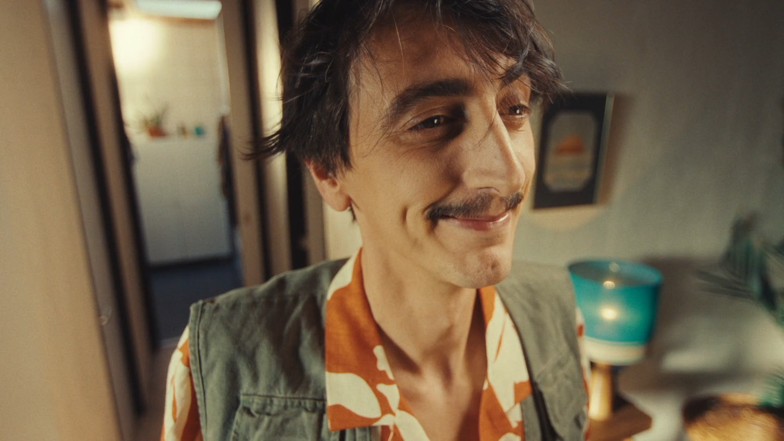 Close-up of a smiling man with dark, wavy hair, mustache, and slight stubble, wearing a patterned shirt and a green jacket, standing in a warmly lit kitchen.