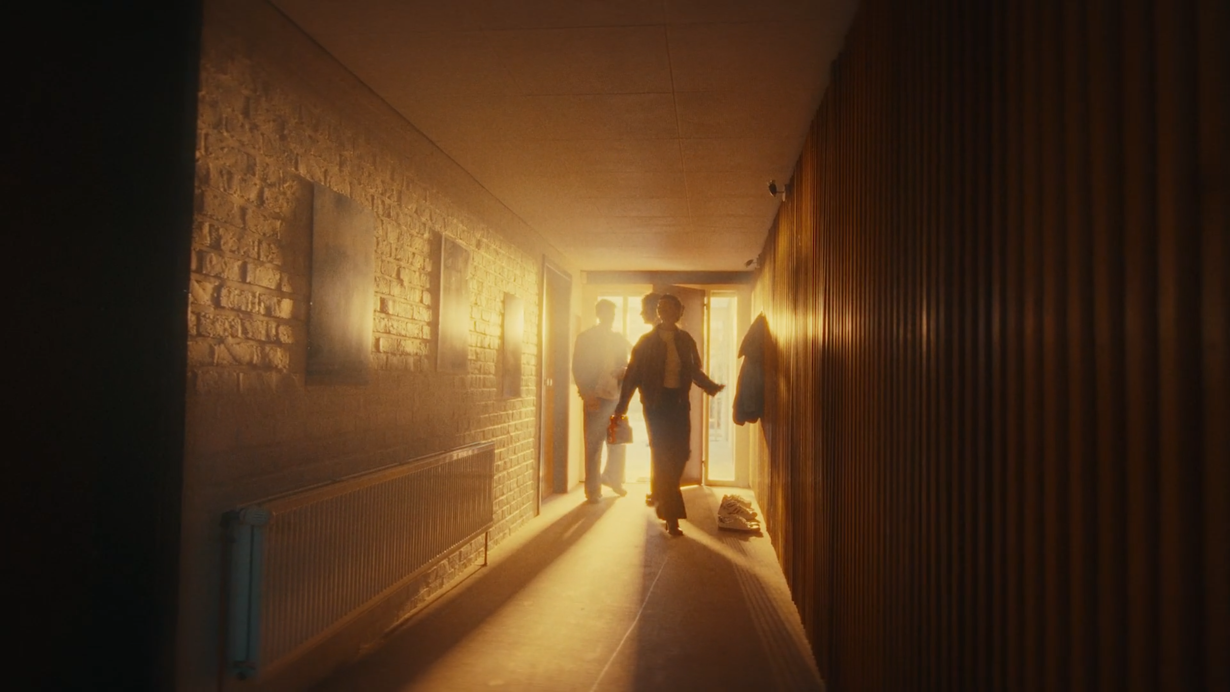 People walking through a brightly lit hallway during sunset, with one person carrying a bag and some shoes on the floor.