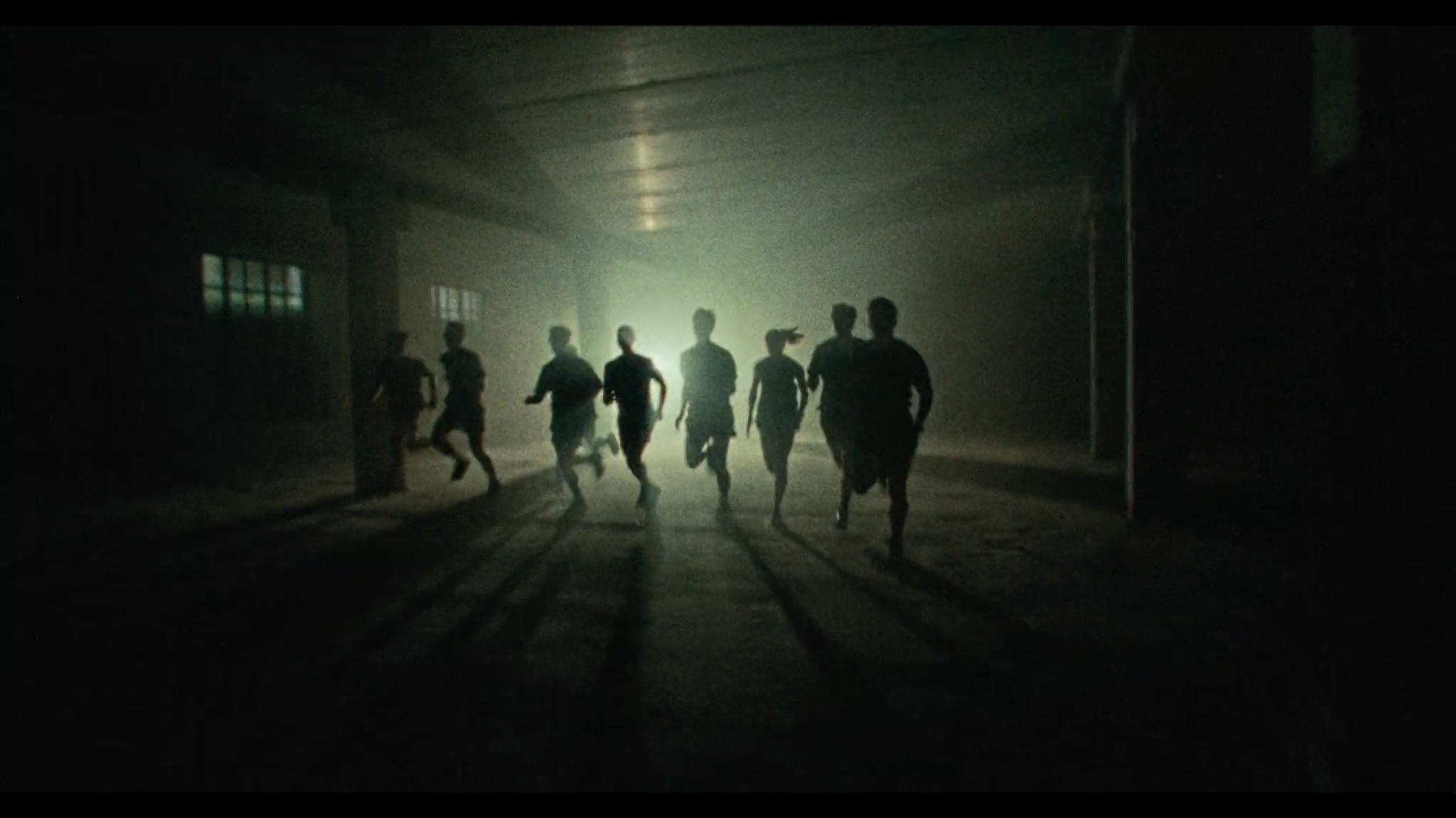 A group of people running through a dark, dimly lit underground space with a bright light source in the background casting long shadows.