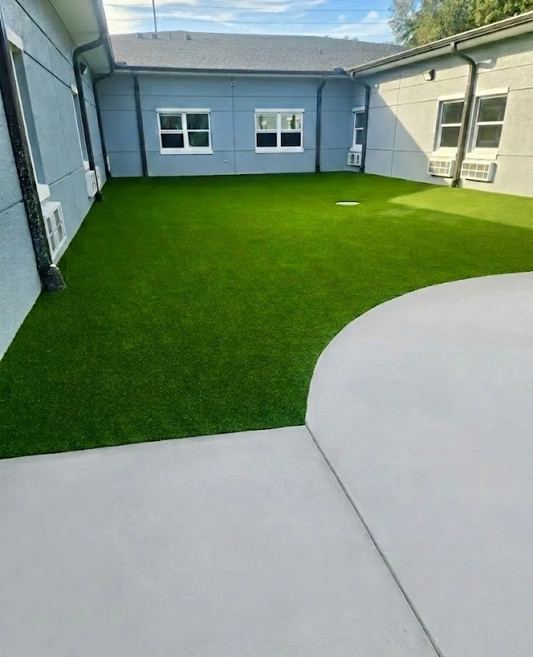 Apartment balcony with artificial grass covering the floor, with white concrete walkway and building walls with several windows and air conditioning units.