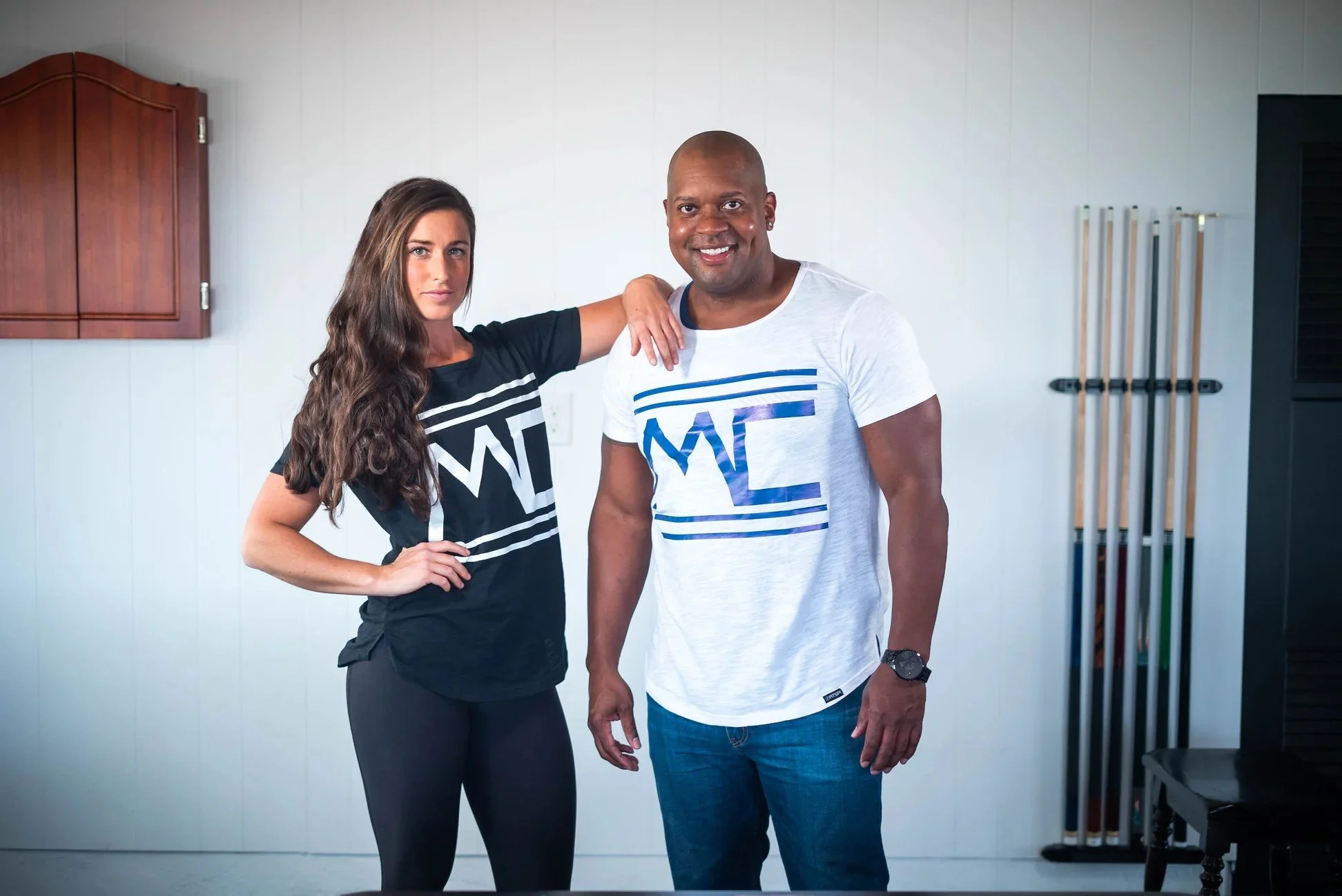 A smiling man with a shaved head, earrings, and a watch, wearing a white t-shirt with blue graphics, sitting against a plain background.