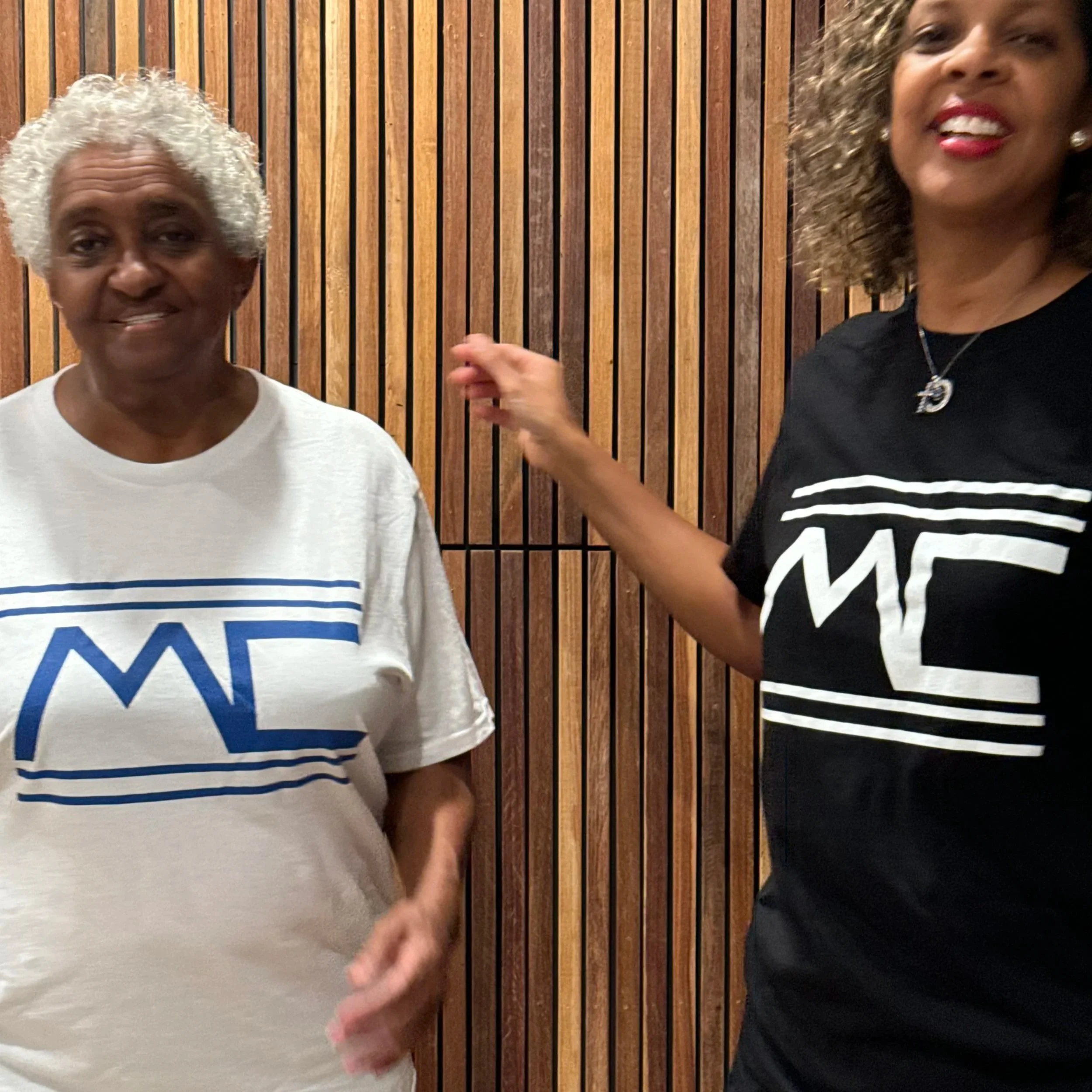 A man wearing a cap and a striped T-shirt smiling and touching his face, with a person in the background playing percussion instruments.