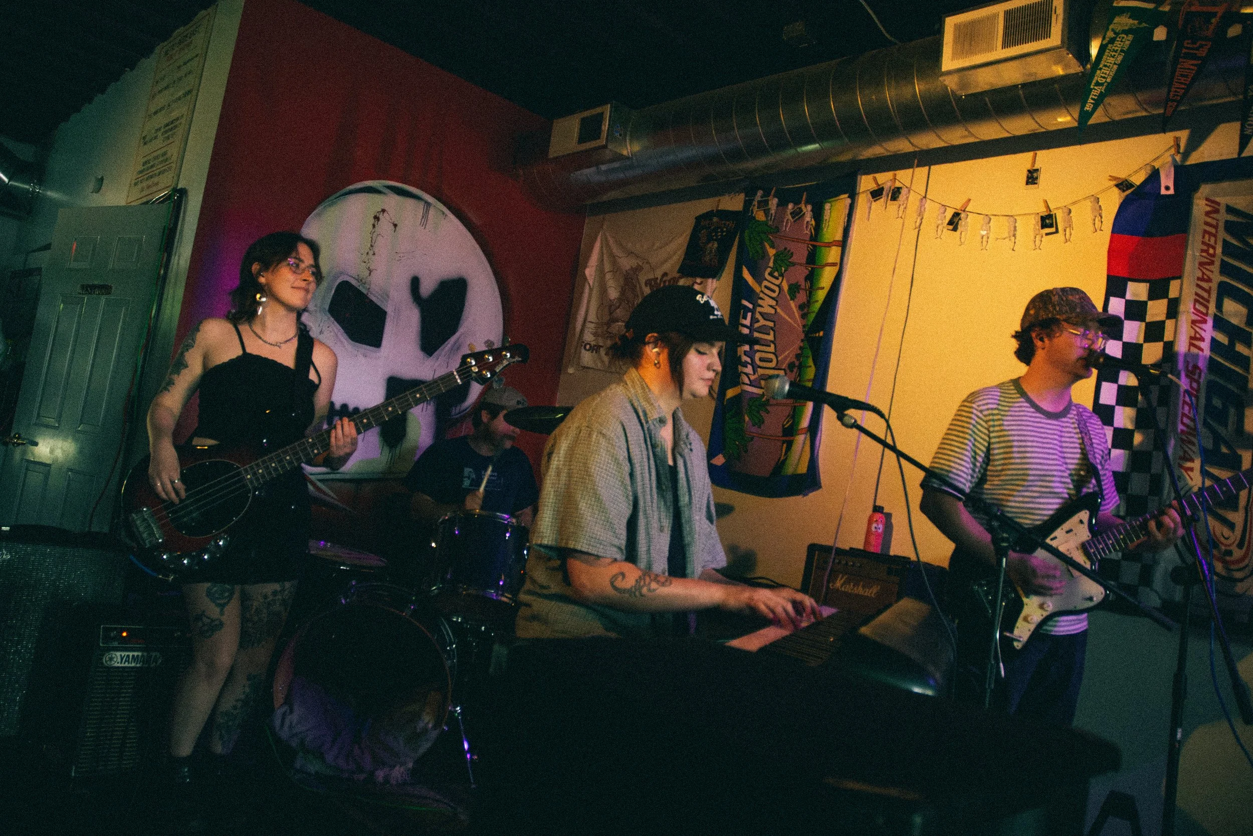 A band of four musicians performing on a stage with colorful banners and a large artwork of a face in the background. One woman plays a bass guitar, a woman sings and plays keyboard, a man plays an electric guitar, and another man drums.