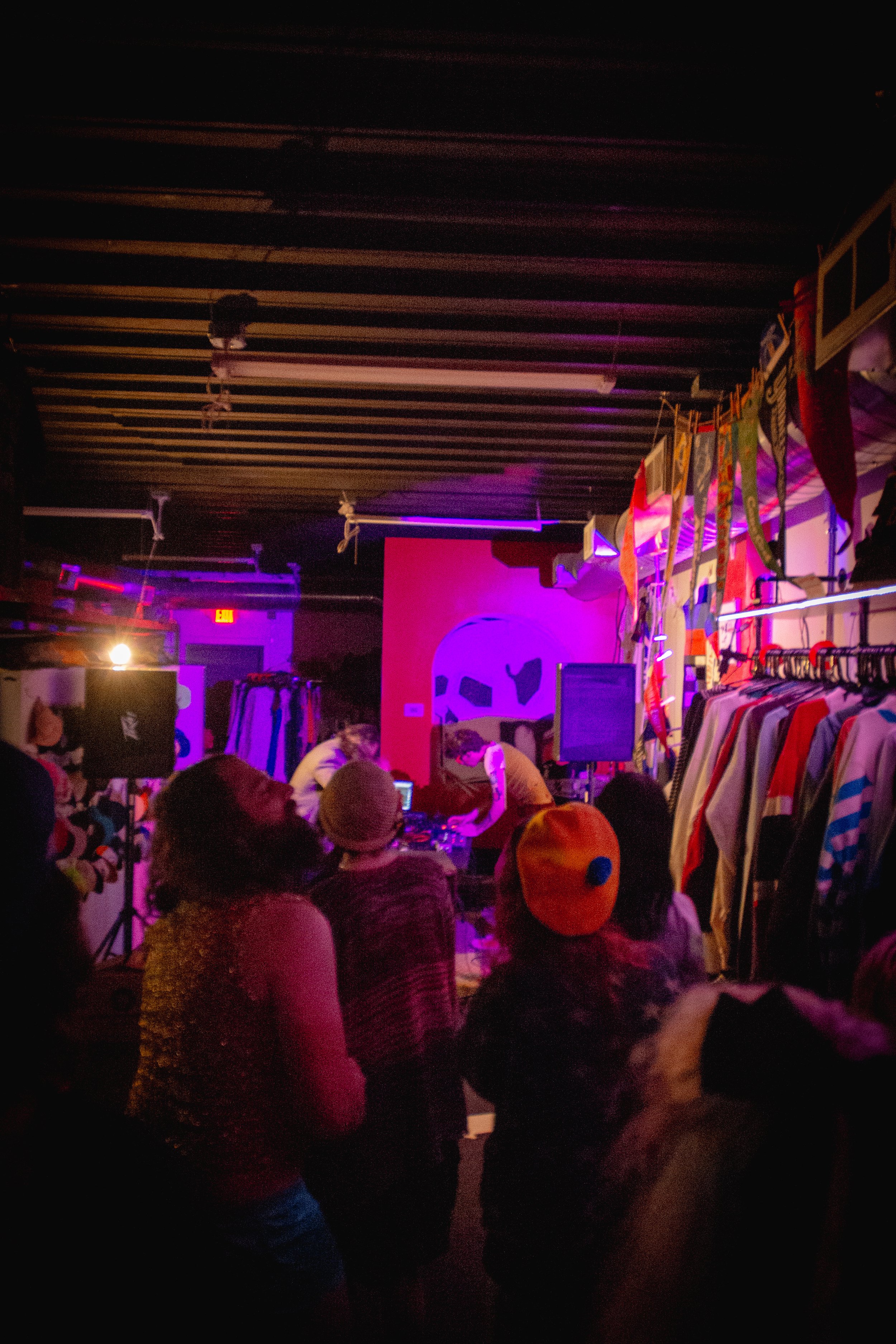 People at a shopping event or market with clothing racks, colorful shirts, and costumes, under purple and pink lighting.