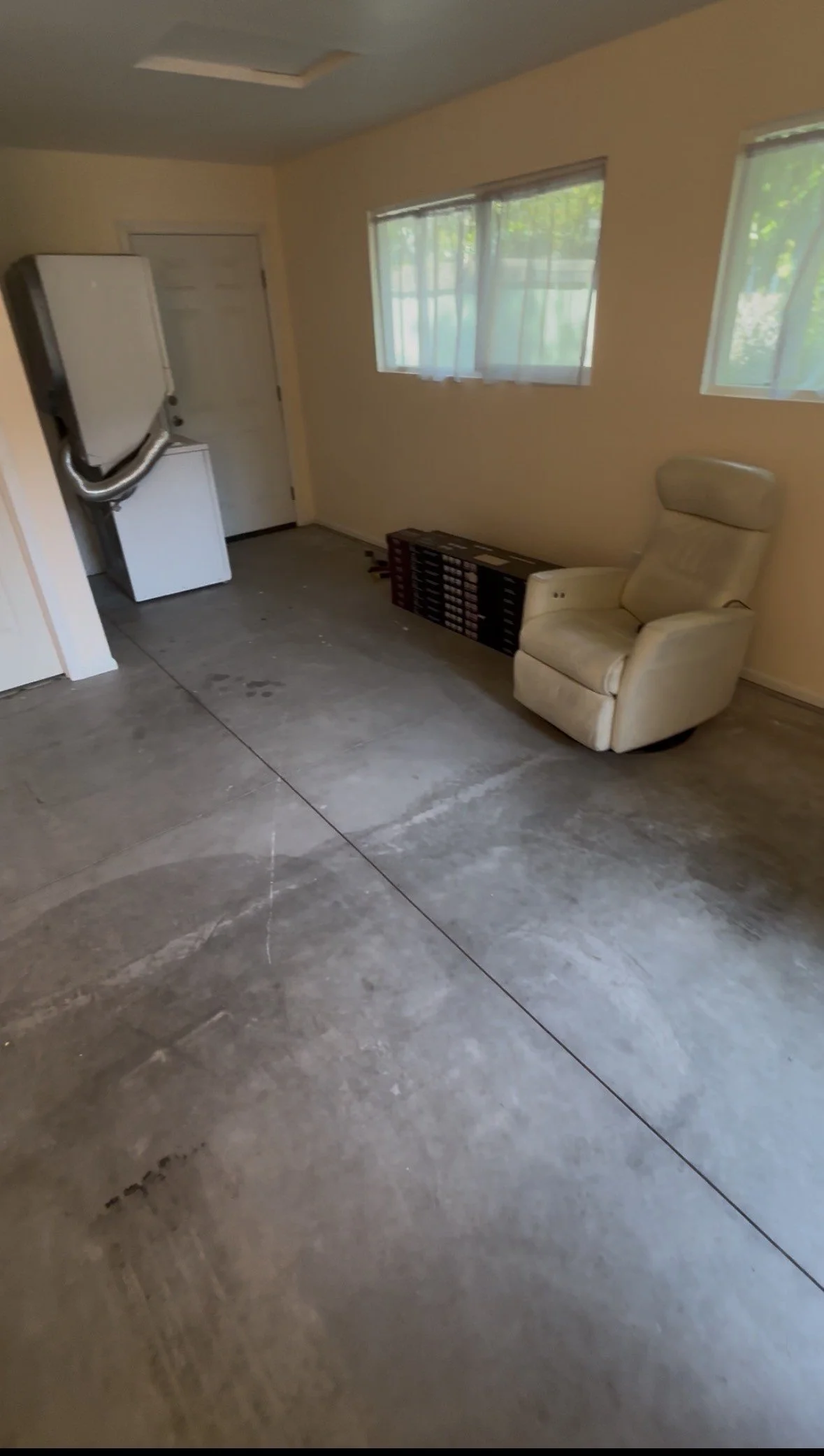 A room with concrete flooring, beige walls, and two windows with sheer curtains. There is a white armchair, some stacked boxes, and a small white appliance near the door. The ceiling has a rectangular vent.