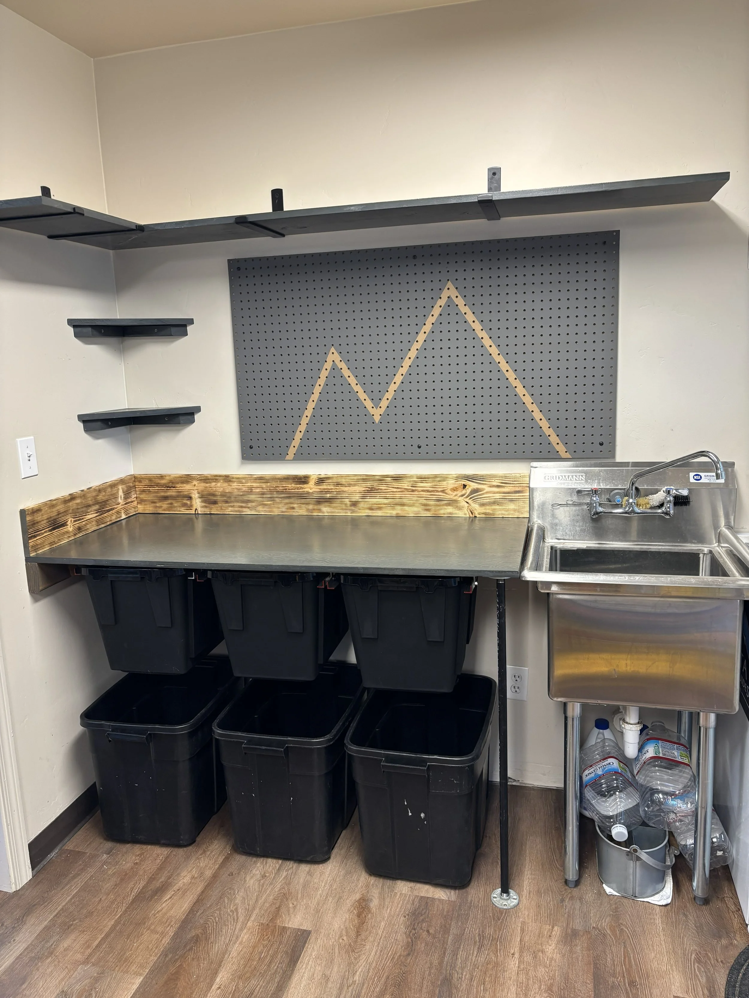 A utility or workshop corner with a stainless steel sink, black storage bins under a wooden countertop, a grey pegboard with a chart, and shelves on the wall.