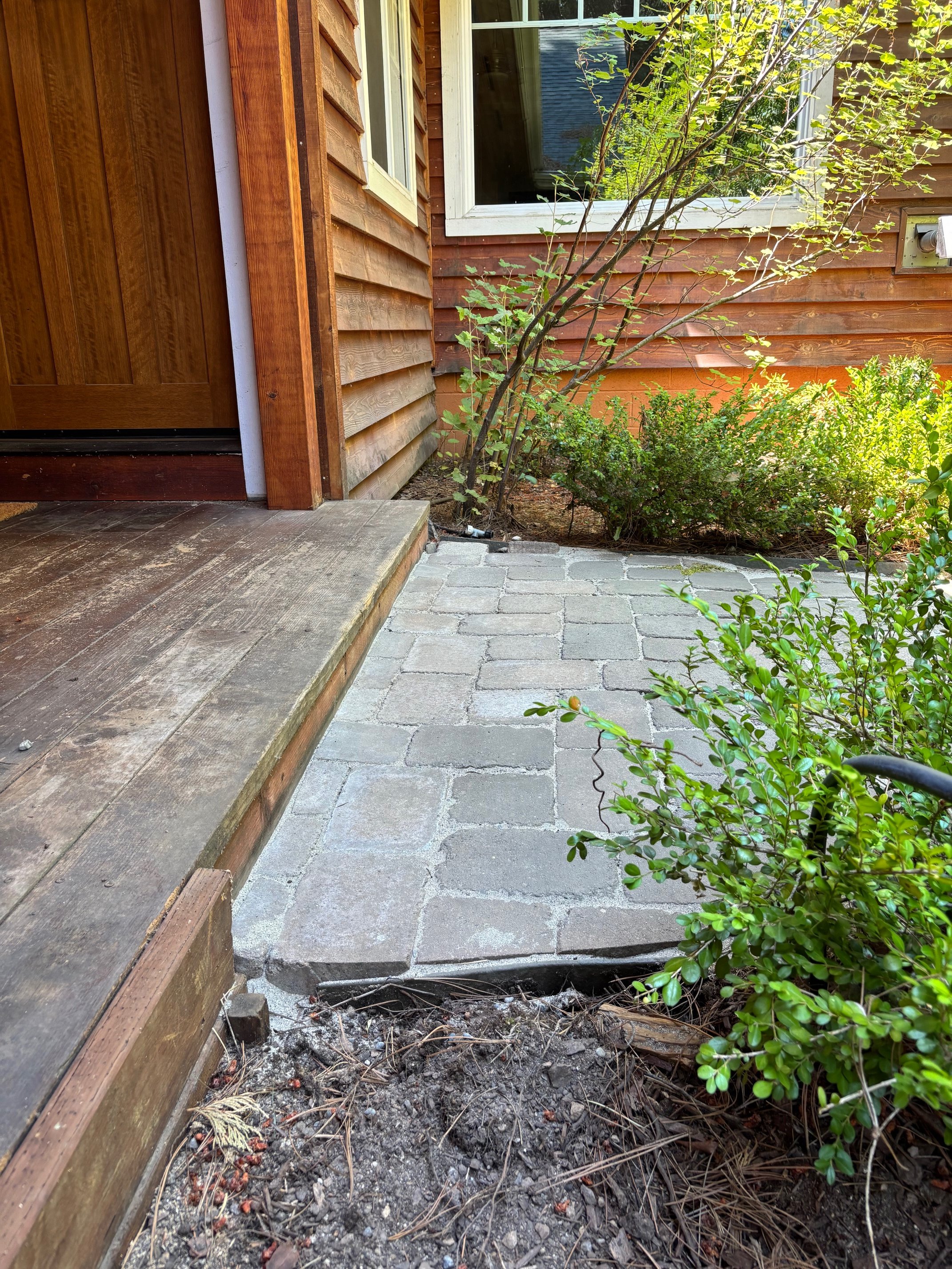 A close-up of a small outdoor patio with a wooden porch and a stone pathway adjacent to a wooden house with a window, surrounded by green shrubs and plants.