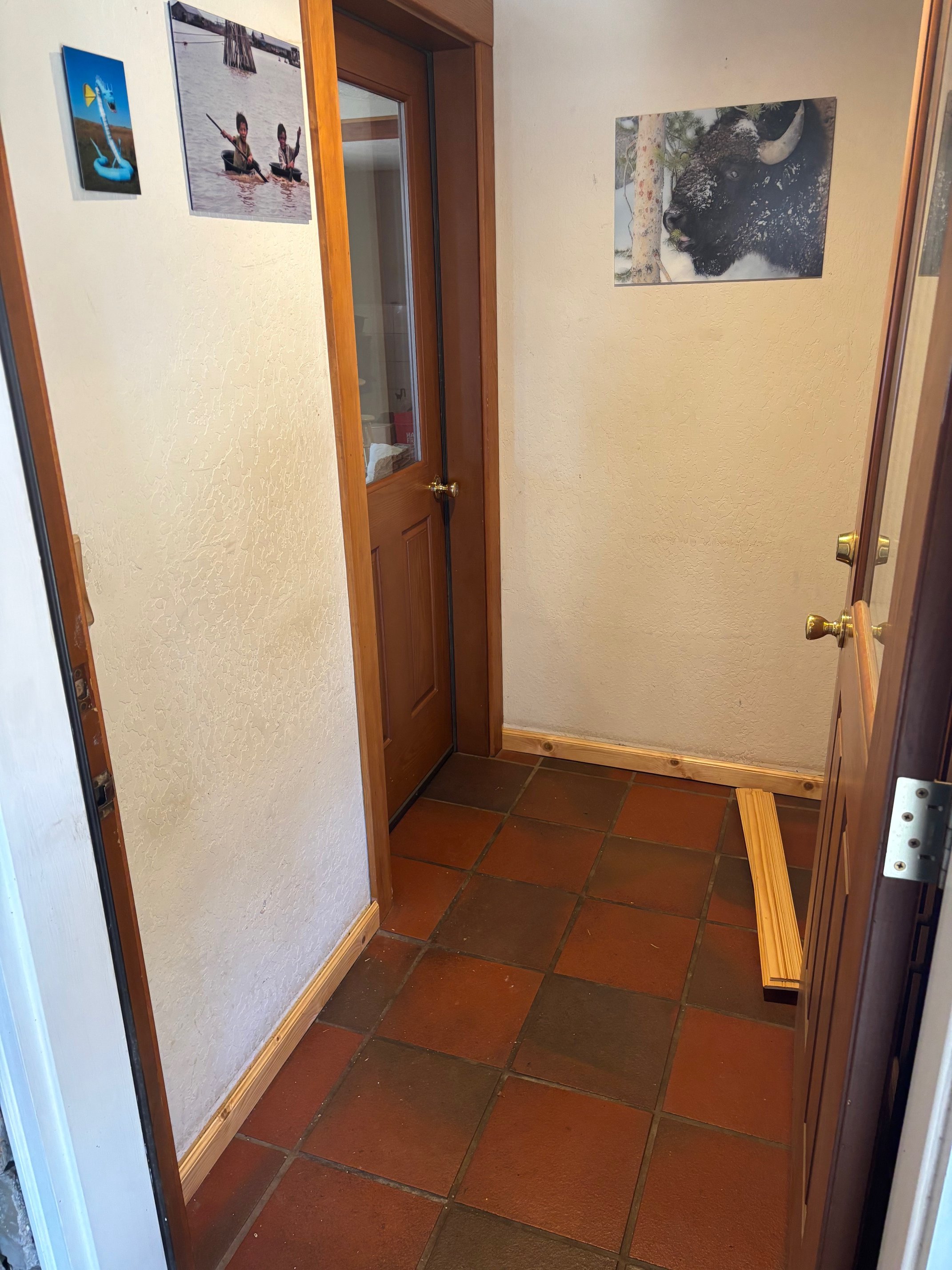 A small entryway with terra cotta tile flooring, wooden trim, a door with a glass panel, and framed photographs on the walls, including a buffalo and children kayaking.