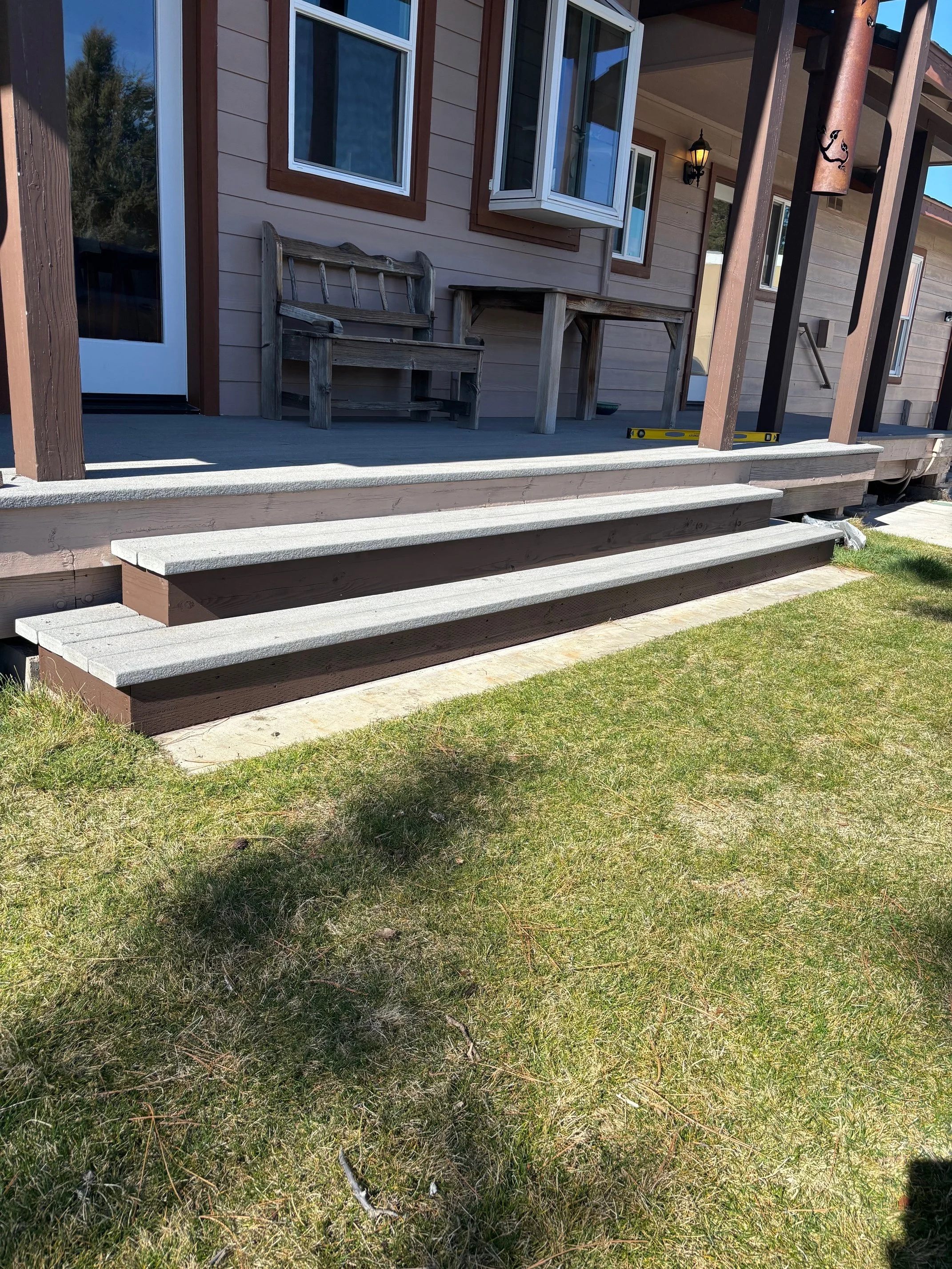 A wooden porch with three steps leading up to a house with beige siding. The porch has light gray flooring and is furnished with a wooden bench, a small table, and a few other items. The house has a window, a door, and an exterior light fixture. There is a grassy lawn in the foreground and some trees in the background.