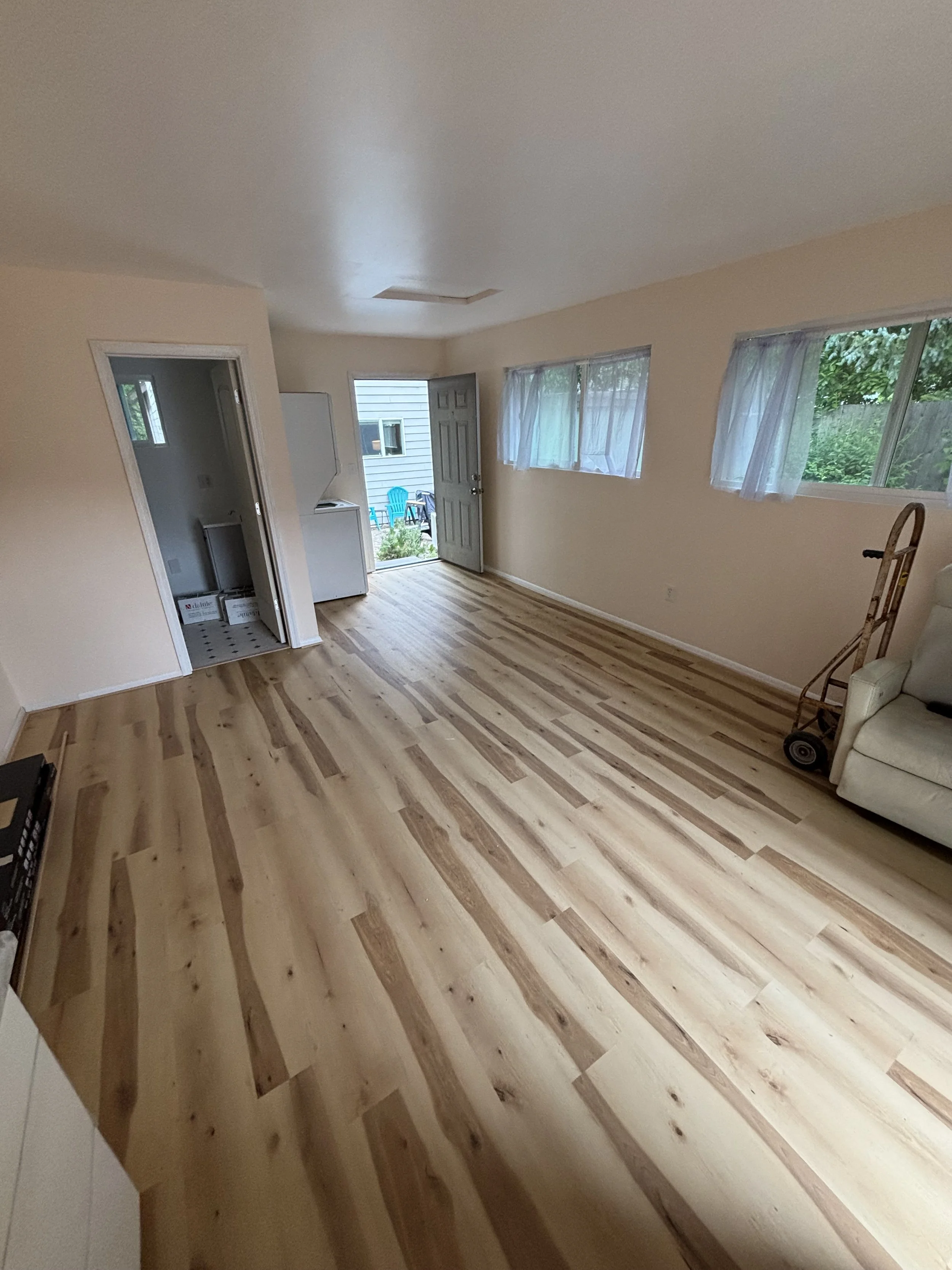 Empty living room with wood flooring, cream walls, three horizontal windows with sheer curtains, open door leading outside with two blue chairs visible, and some furniture and items in the room.