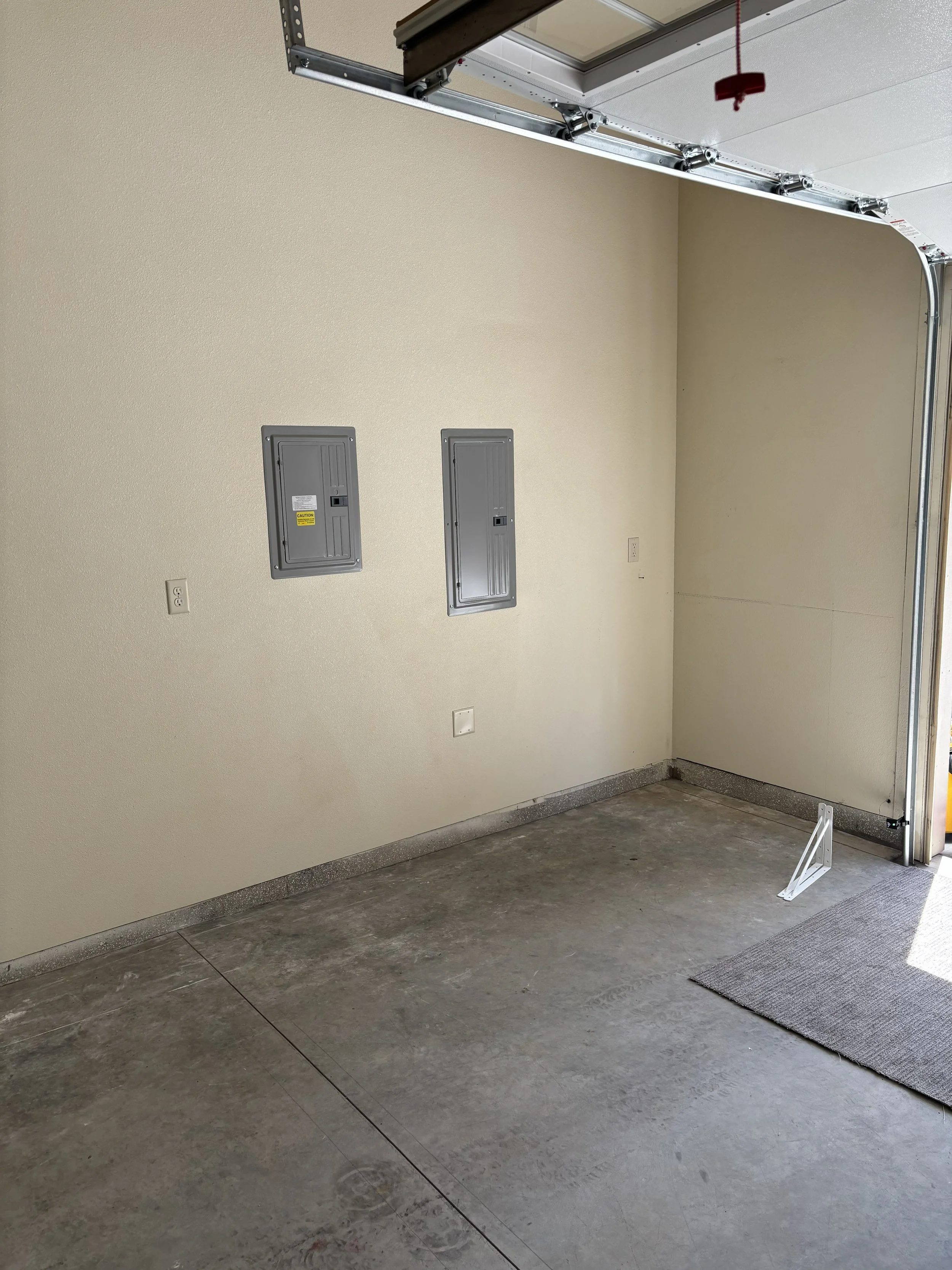 Empty garage with a closed garage door, beige painted walls, two electrical panels on the wall, and a gray doormat on a concrete floor.