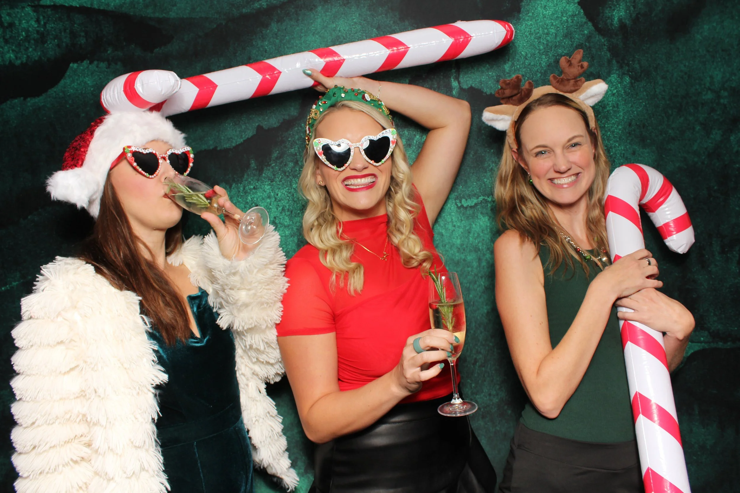 Three women in Christmas-themed costumes celebrating with drinks and festive props, including Santa hat, reindeer antlers, and holiday glasses, against a festive green photo booth backdrop