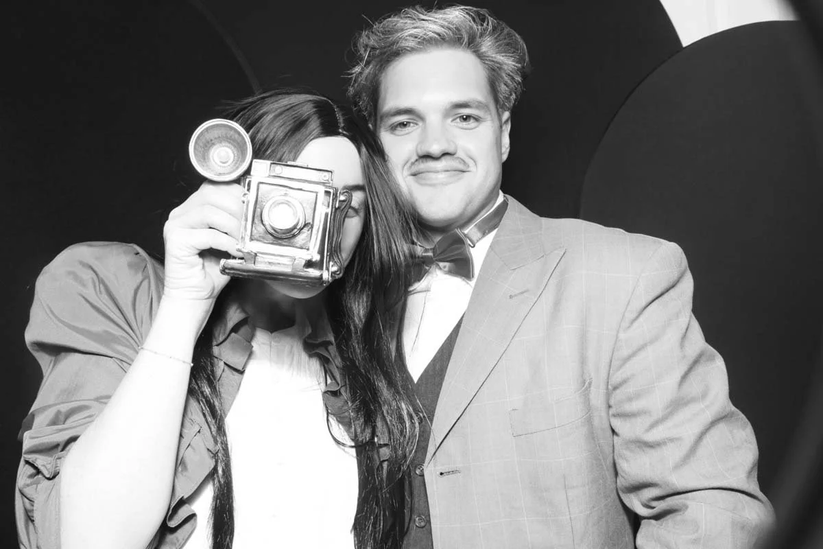 A man and woman share a playful moment in a vintage black and white photo booth, showcasing their smiles and connection